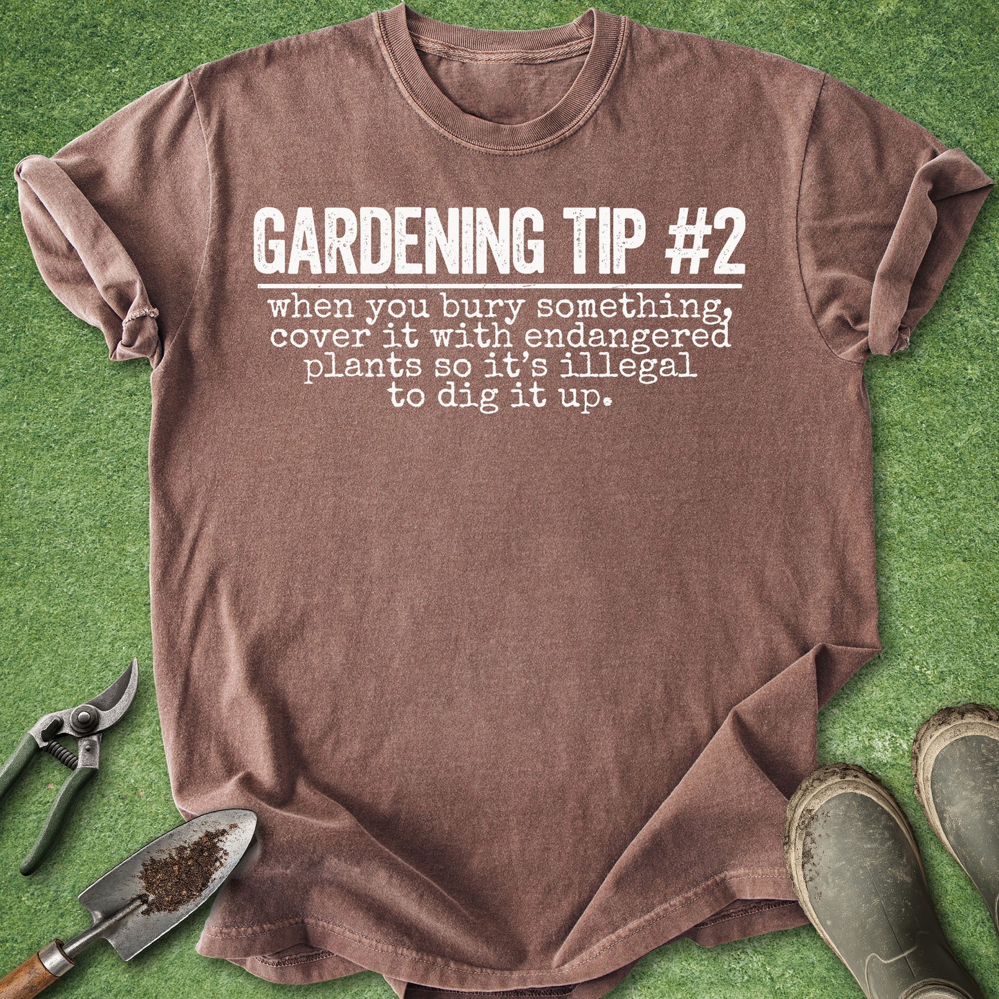 A brown short-sleeve t-shirt with white printed text reading 'GARDENING TIP #2 when you bury something, cover it with endangered plants so it's illegal to dig it up.' laid flat on green grass-like background with gardening props including pruning shears, trowel, and boots around it.