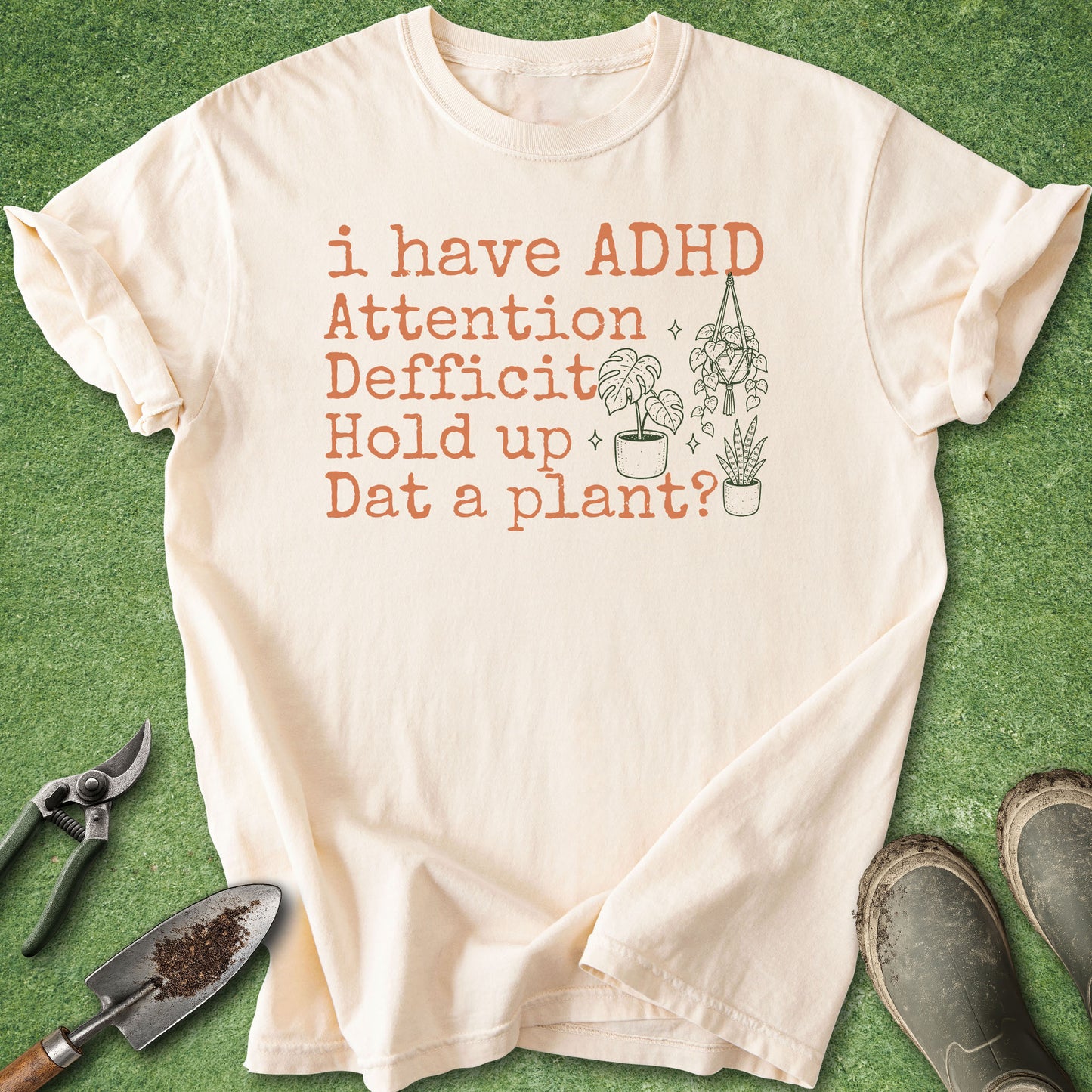 A cream-colored short-sleeve t-shirt with a humorous graphic print reading 'i have ADHD Attention Deficit Hold up Dat a plant?' in terracotta orange text, accompanied by a simple line drawing of potted plants. The shirt is laid flat on a green grass-like surface, styled with gardening tools (trowel, pruning shears) and work boots.