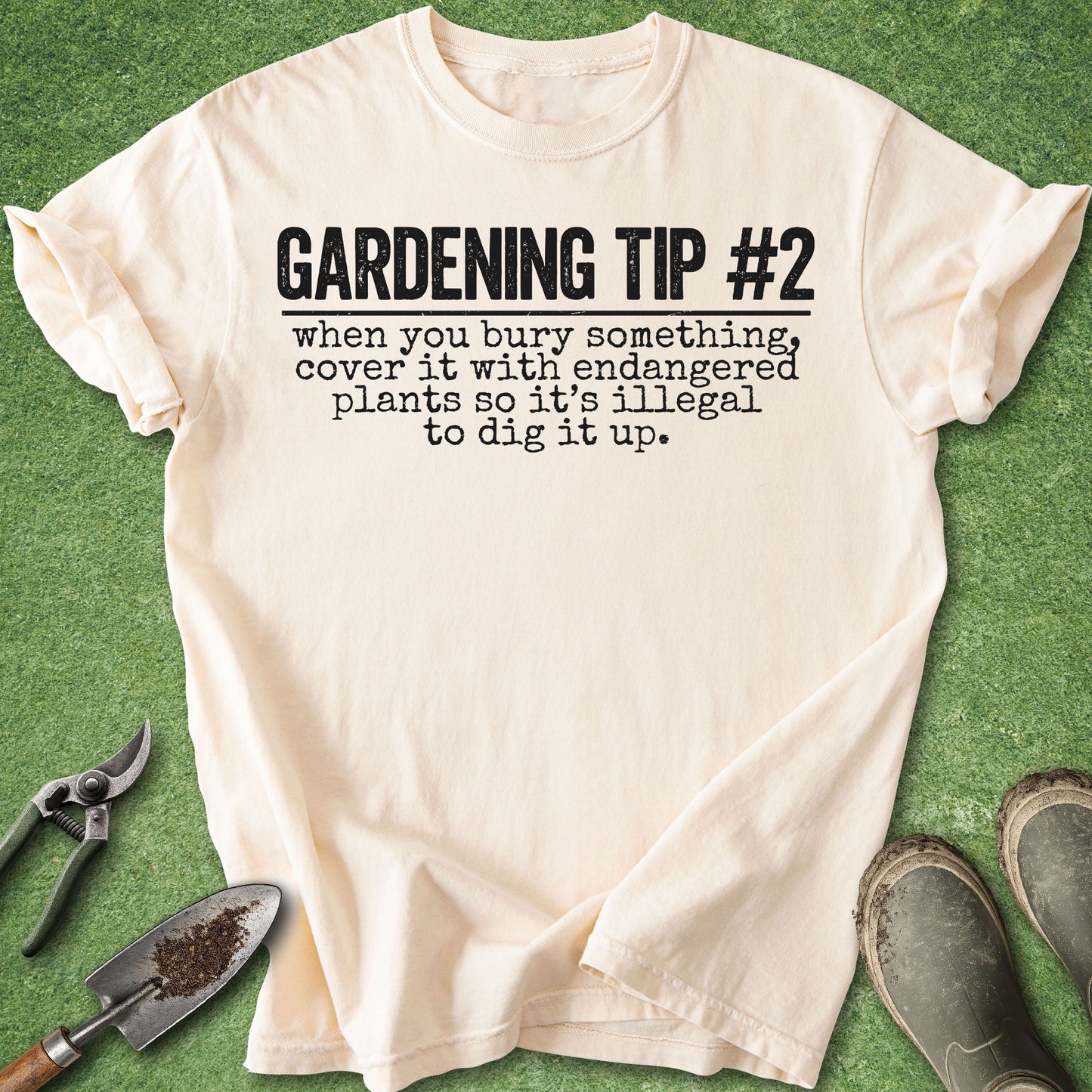 A cream-colored short-sleeve t-shirt with black printed text reading 'GARDENING TIP #2 when you bury something, cover it with endangered plants so it's illegal to dig it up.' The shirt is laid flat on green artificial grass with gardening props including pruning shears, a trowel, and boots around it.