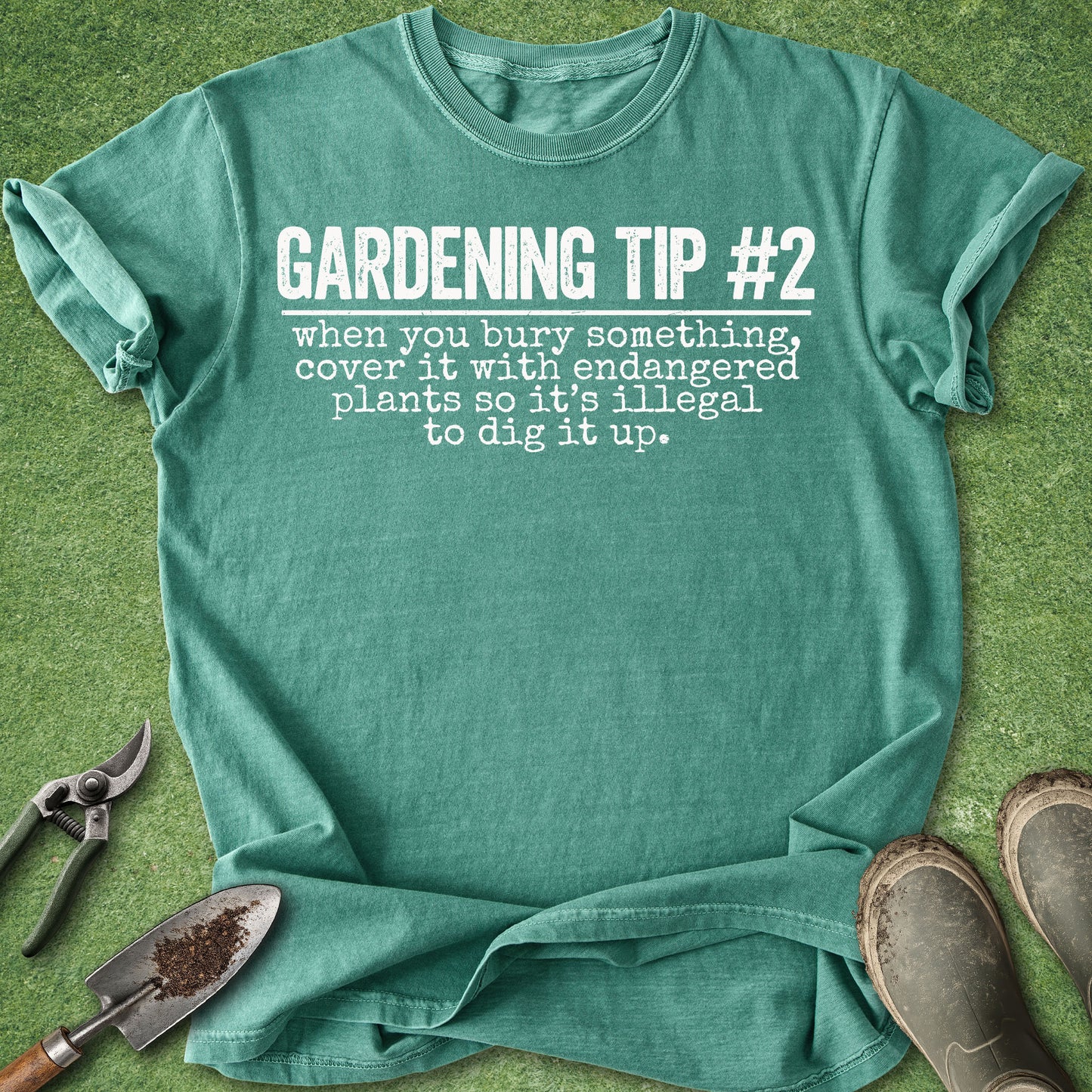 A teal green graphic t-shirt laid flat on green grass-like background, with gardening tools and boots around it. The shirt reads 'GARDENING TIP #2 when you bury something, cover it with endangered plants so it's illegal to dig it up.