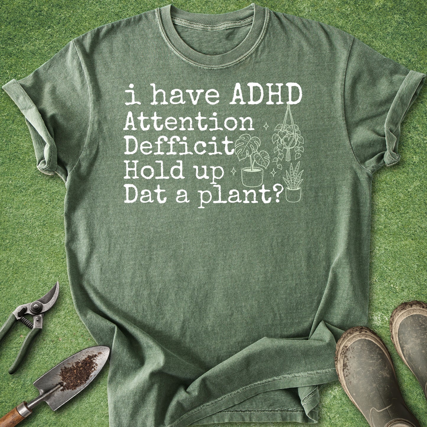 An olive green graphic t-shirt laid flat on a green grass-like surface, surrounded by gardening props including pruning shears, a trowel, and work boots. The shirt features white distressed text that reads 'i have ADHD Attention Deficit Hold up Dat a plant?' with simple line drawings of potted plants.