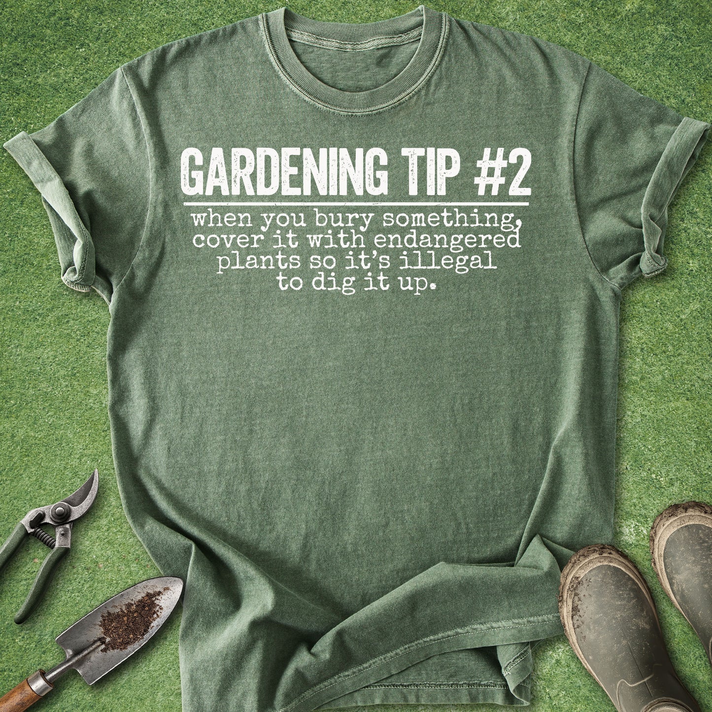 An olive green graphic t-shirt laid flat on a green grass-like surface, with gardening tools and boots around it. The shirt reads, 'GARDENING TIP #2 when you bury something, cover it with endangered plants so it's illegal to dig it up.