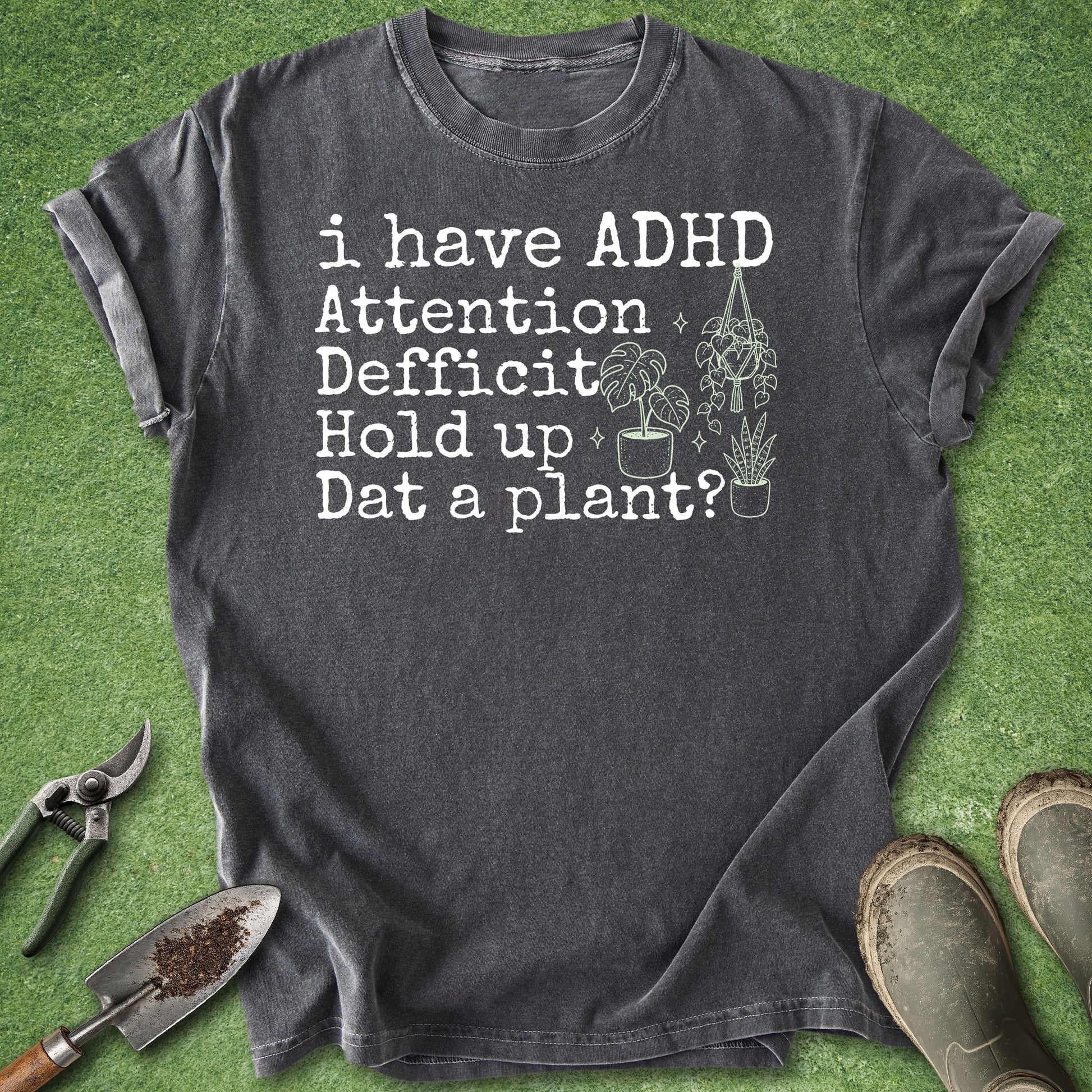 A dark charcoal gray graphic t-shirt laid flat on a green grass-like surface, with gardening tools and boots around it. The shirt has white text reading 'i have ADHD Attention Deficit Hold up Dat a plant?' with small plant illustrations.