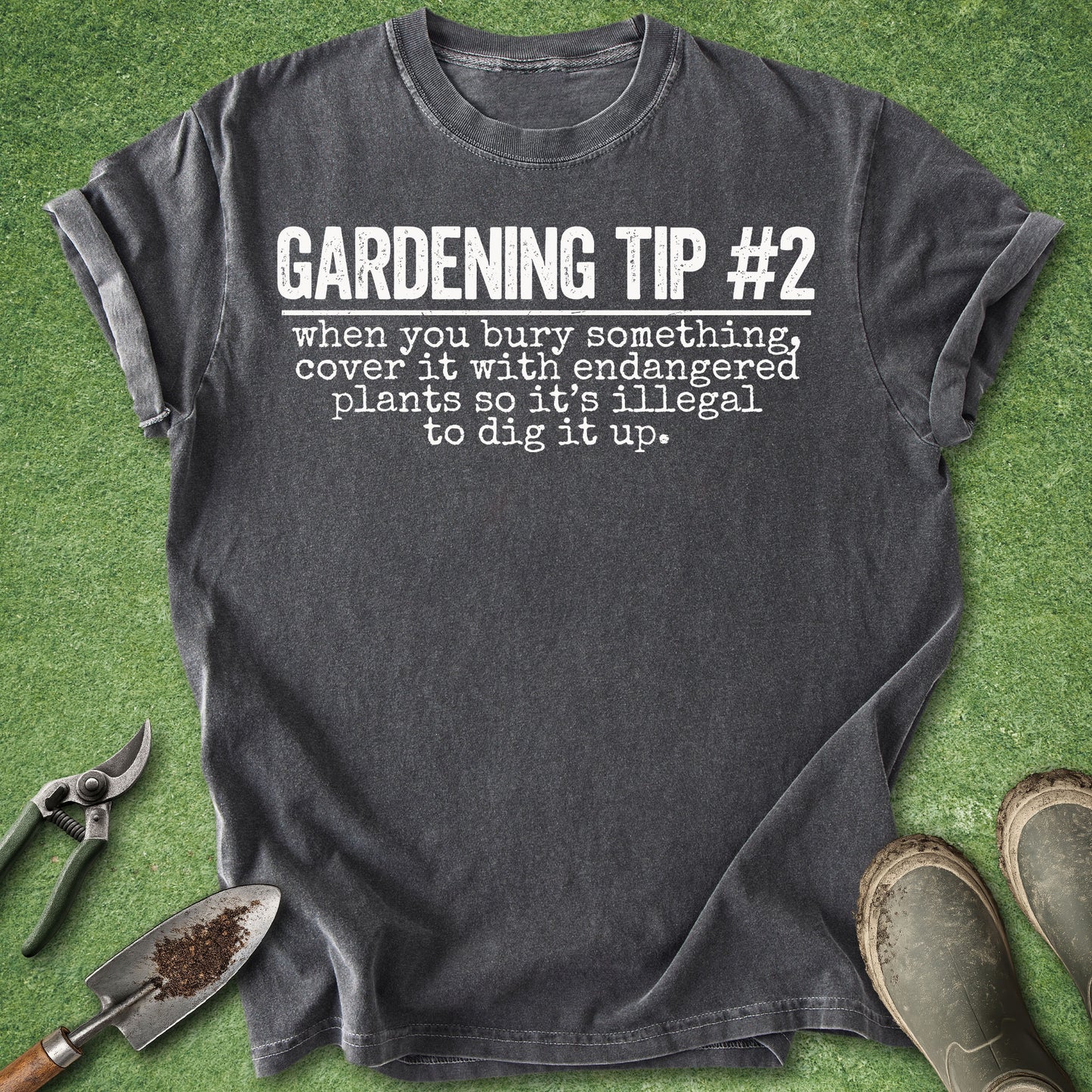 A dark charcoal gray graphic t-shirt laid flat on artificial green grass, styled with gardening tools (trowel, pruning shears) and muddy boots. The t-shirt features the text 'GARDENING TIP #2 when you bury something, cover it with endangered plants so it's illegal to dig it up.