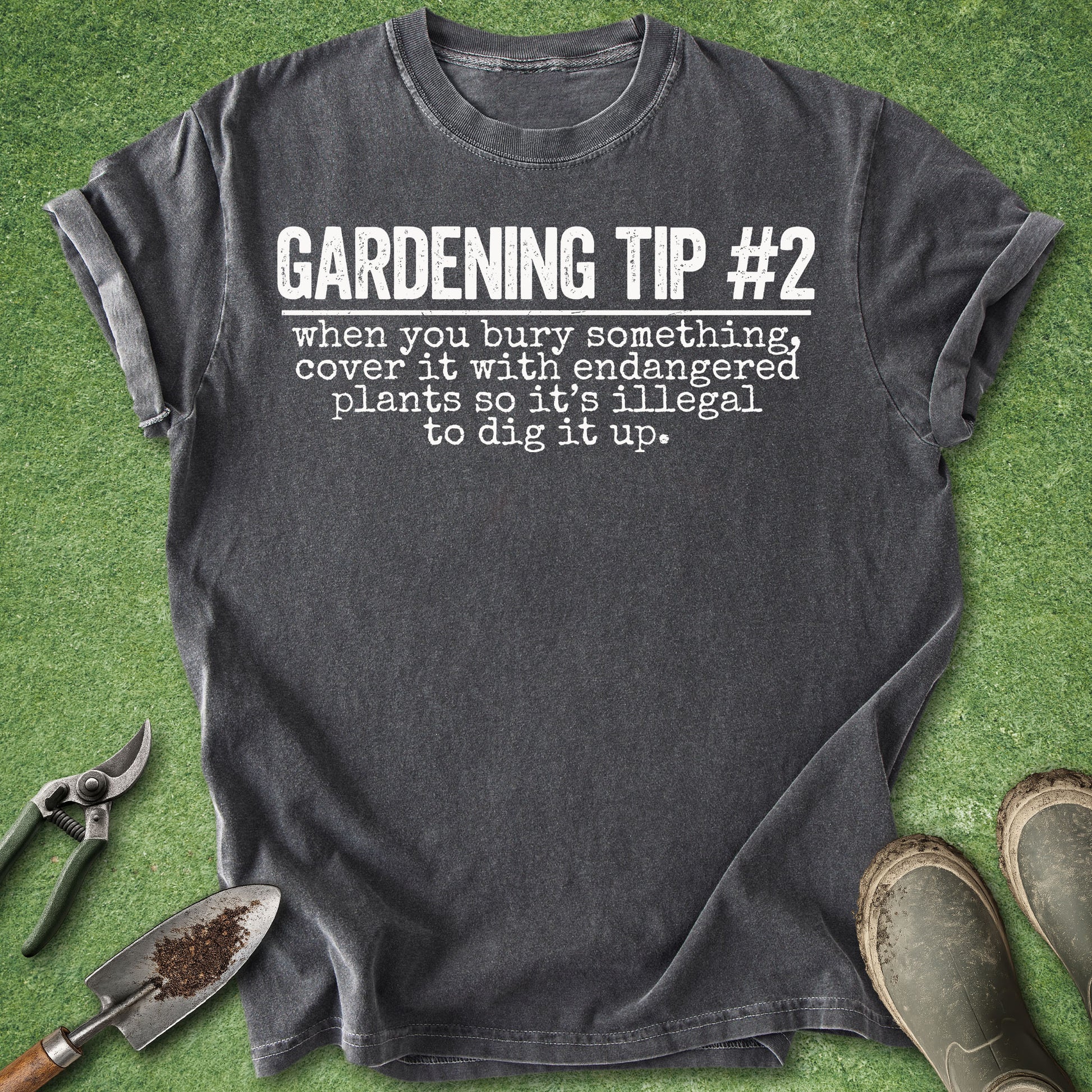 A dark charcoal gray graphic t-shirt laid flat on artificial green grass, styled with gardening tools (trowel, pruning shears) and muddy boots. The t-shirt features the text 'GARDENING TIP #2 when you bury something, cover it with endangered plants so it's illegal to dig it up.