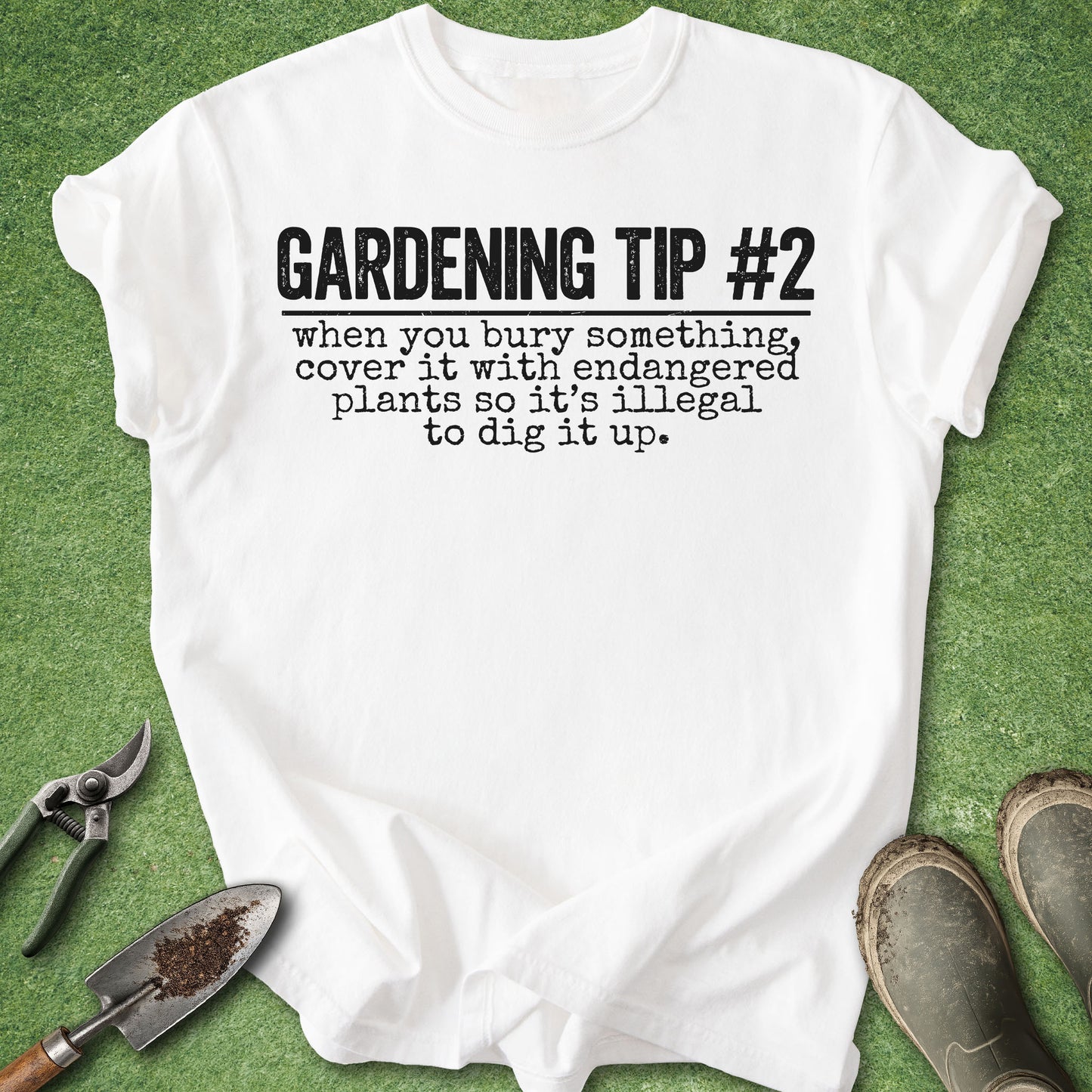 A white graphic t-shirt laid flat on green artificial grass, with gardening tools and boots around it. The shirt reads 'GARDENING TIP #2 when you bury something, cover it with endangered plants so it's illegal to dig it up.