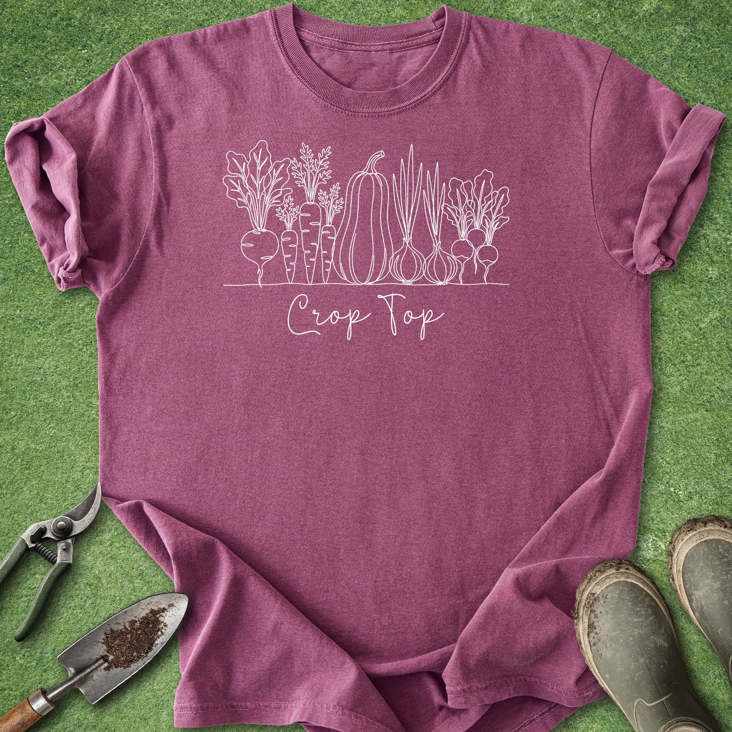 A heathered mauve short-sleeve t-shirt with a white line art graphic of various vegetables and the text 'Crop Top' printed on the chest. The shirt is laid flat on green grass with gardening props, including a trowel, pruning shears, and boots.