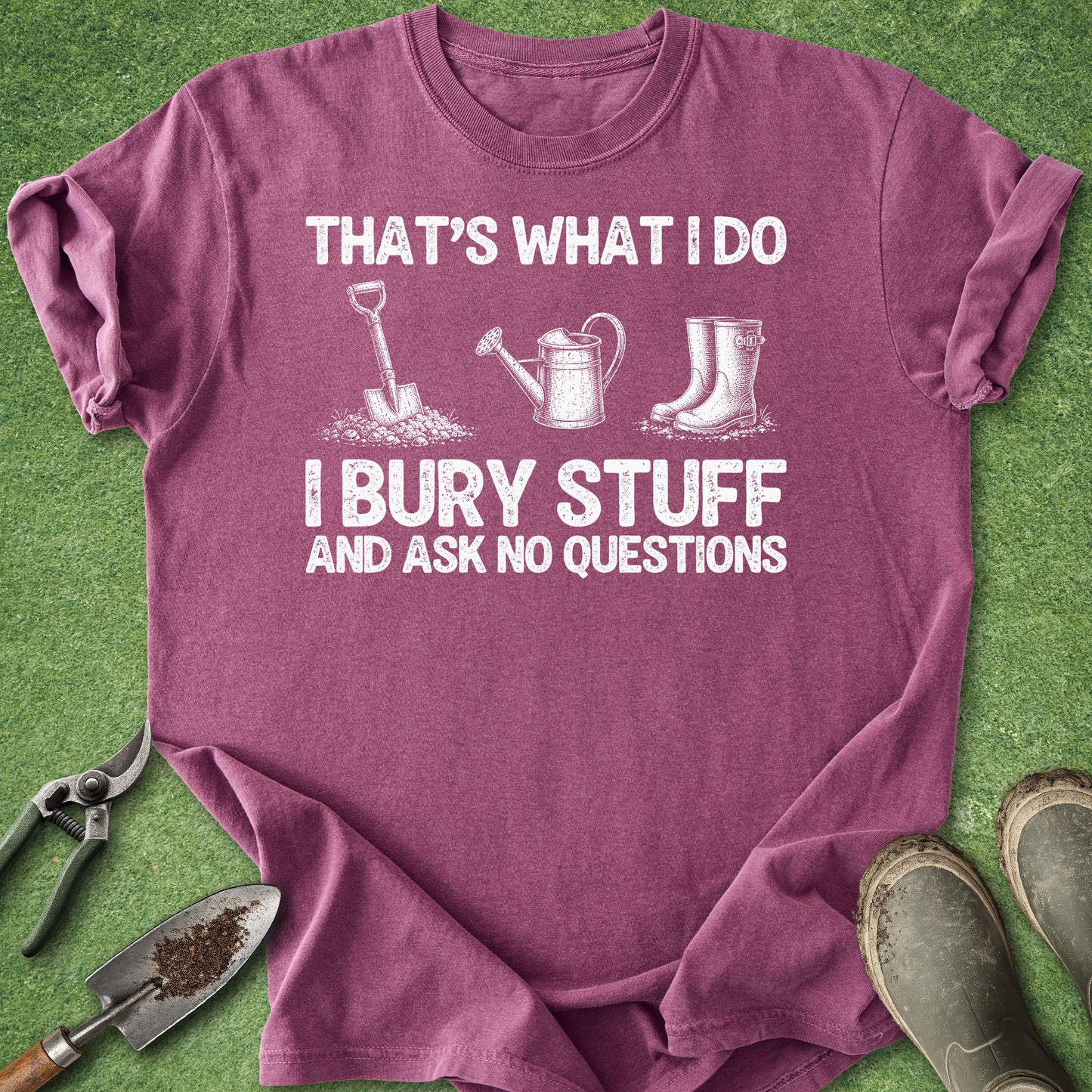 A heathered purple short-sleeve t-shirt with a white graphic print that reads 'THAT'S WHAT I DO I BURY STUFF AND ASK NO QUESTIONS' with illustrations of a shovel, watering can, and rain boots. The shirt is laid flat on green grass-like artificial turf with gardening tools and boots around it.
