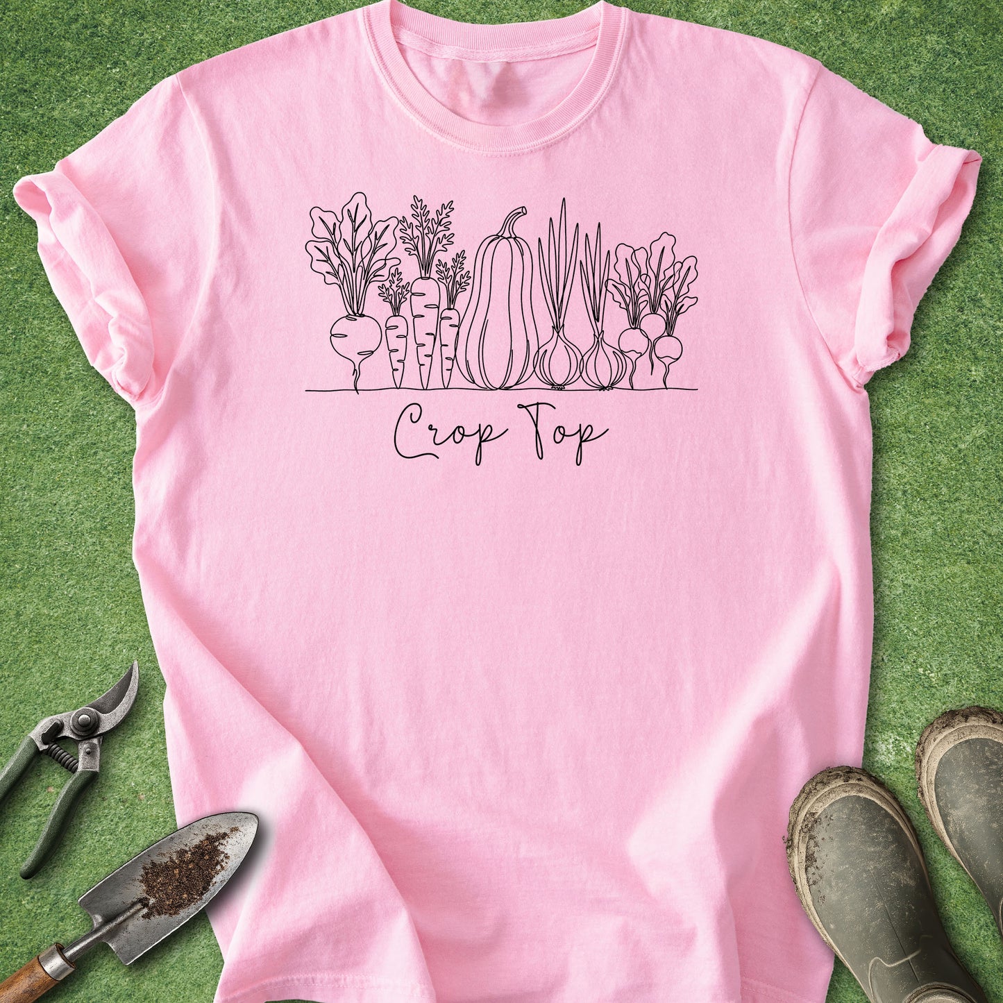 A pink short-sleeve T-shirt laid flat on green grass, featuring a black line-art vegetable illustration and the words 'Crop Top' across the chest. Gardening tools and boots are placed around the shirt as styling props.