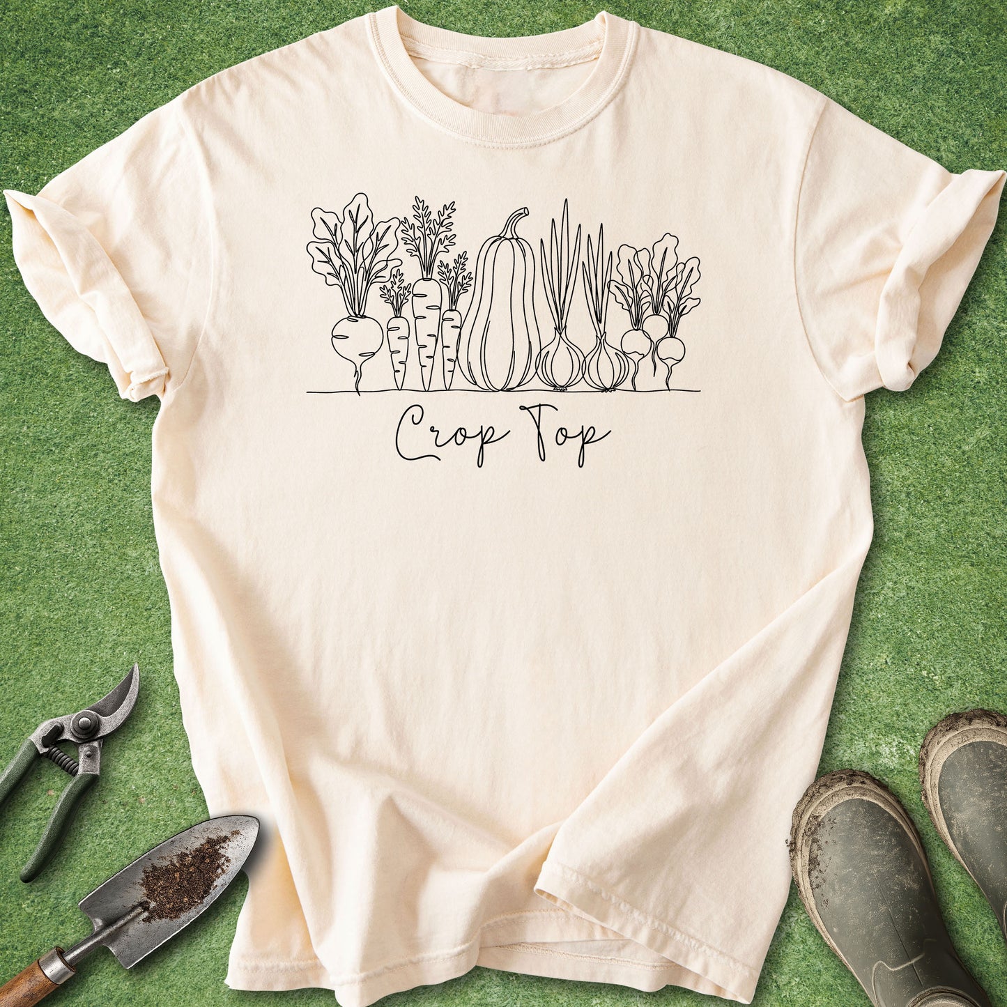A cream-colored short-sleeve t-shirt with a black line art graphic of various vegetables and the text 'Crop Top' underneath, laid flat on a green grass-like background with gardening props including a trowel, pruning shears, and boots.