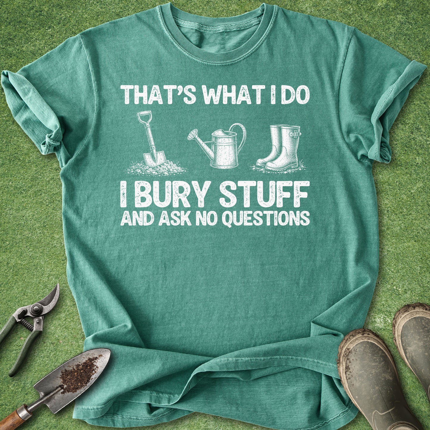 A teal green graphic t-shirt laid flat on a green grass-like surface, surrounded by gardening props including pruning shears, a trowel, and boots. The shirt features a white printed graphic with the text 'THAT'S WHAT I DO I BURY STUFF AND ASK NO QUESTIONS' and simple illustrations of a shovel, watering can, and rain boots.