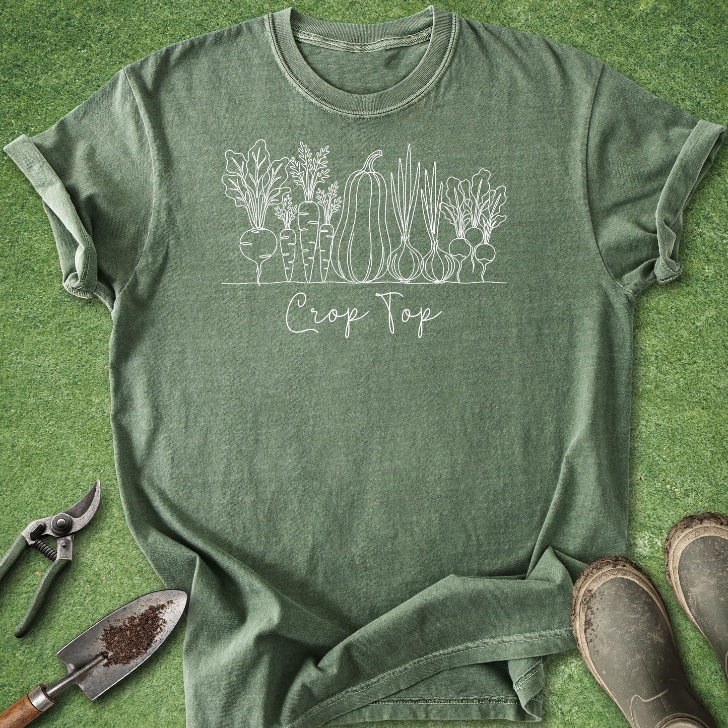 An olive green heathered t-shirt with a white line art graphic of vegetables and the text 'Crop Top', laid flat on a green grass-like background with gardening props (trowel, pruning shears, boots) around it.