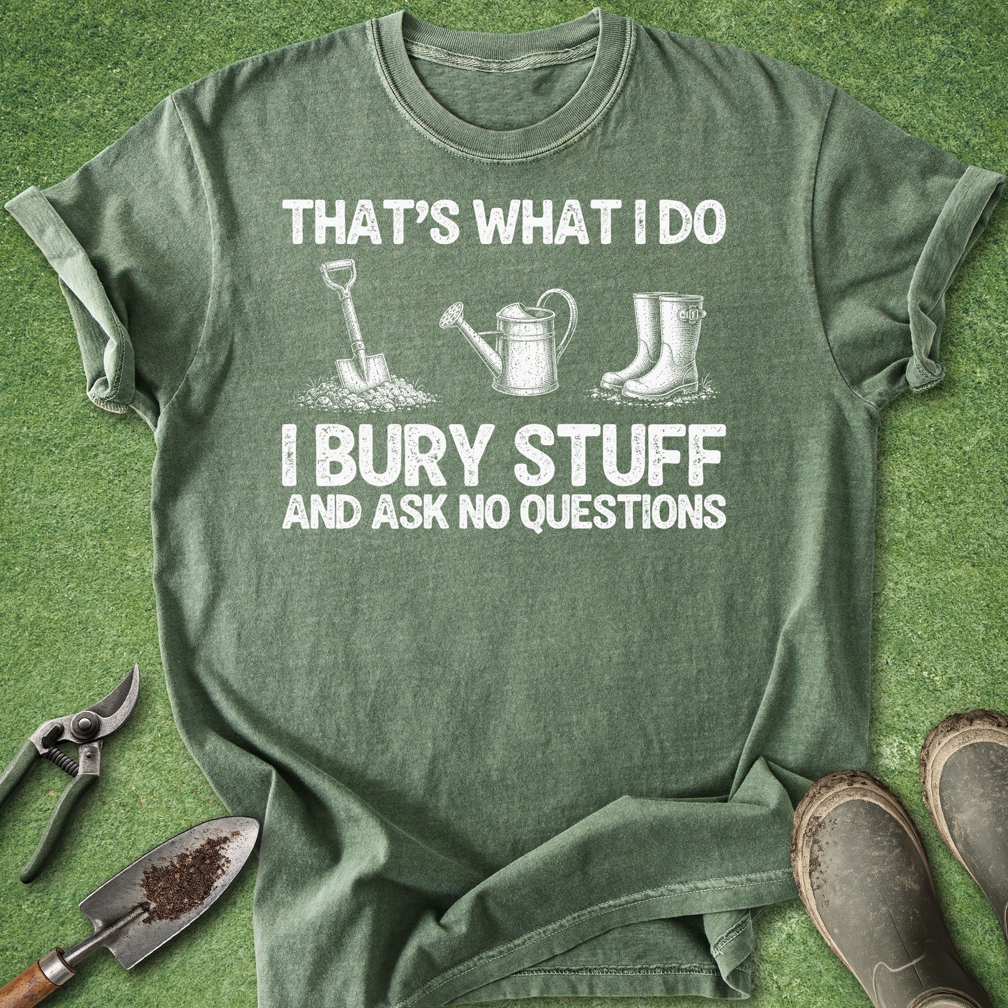 An olive green graphic t-shirt laid flat on a green grass-like surface, surrounded by gardening props including pruning shears, a trowel, and boots. The shirt features a white printed graphic with the text 'THAT'S WHAT I DO I BURY STUFF AND ASK NO QUESTIONS' and simple illustrations of a shovel, watering can, and rain boots.