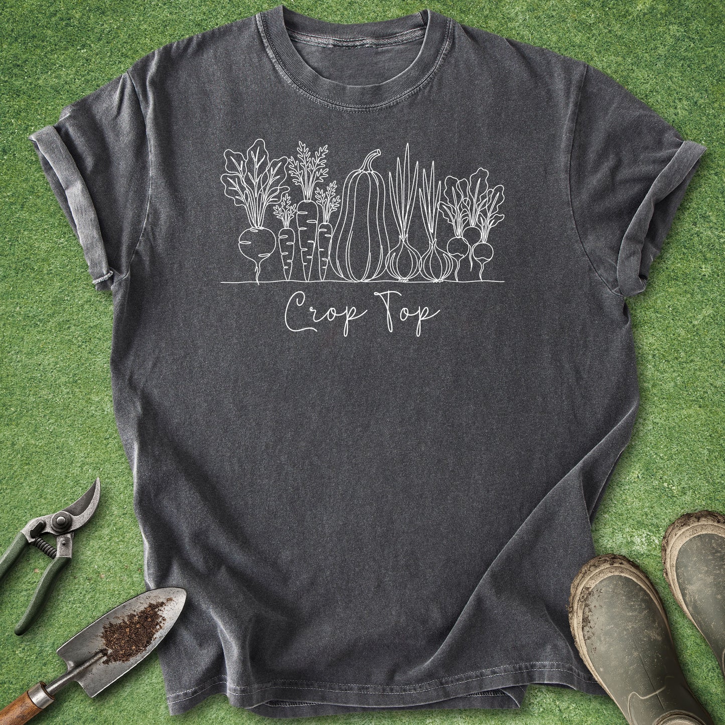 A dark charcoal gray t-shirt with a white line art graphic of vegetables and the text 'Crop Top', laid flat on a green grass-like background with gardening tools and boots nearby.