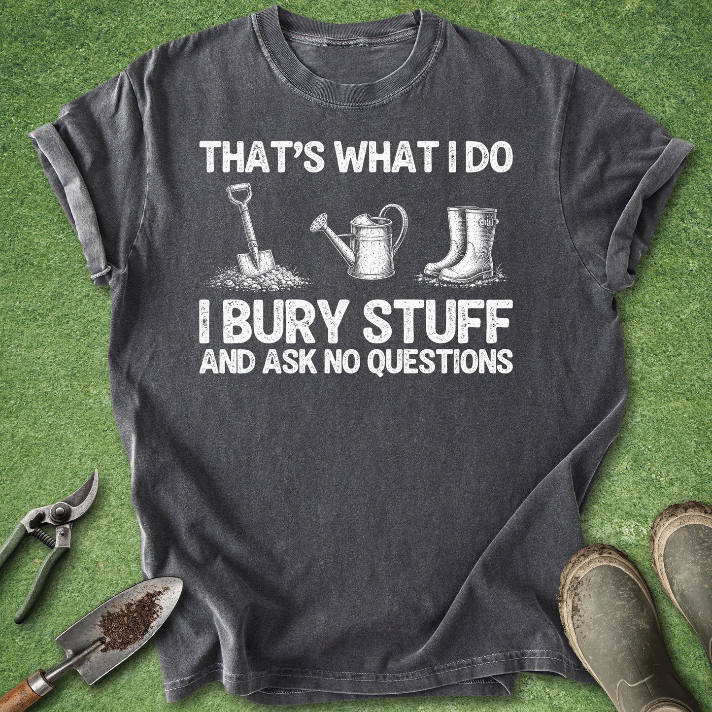 A dark gray graphic t-shirt laid flat on green grass, surrounded by gardening props including pruning shears, a trowel, a watering can, and boots. The shirt features white text reading 'THAT'S WHAT I DO I BURY STUFF AND ASK NO QUESTIONS' with corresponding gardening tool illustrations.