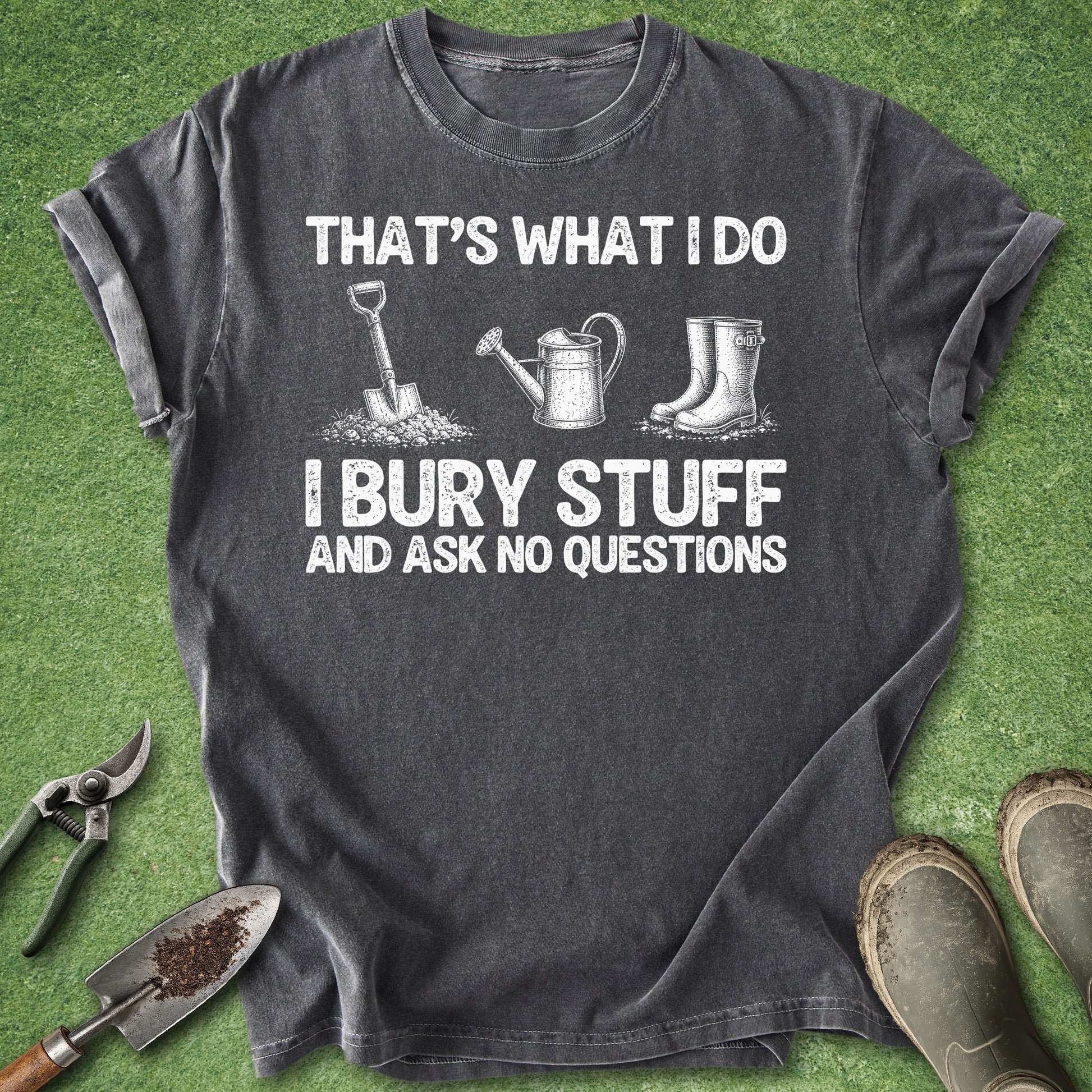 A dark gray graphic t-shirt laid flat on green grass, surrounded by gardening props including pruning shears, a trowel, a watering can, and boots. The shirt features white text reading 'THAT'S WHAT I DO I BURY STUFF AND ASK NO QUESTIONS' with corresponding gardening tool illustrations.