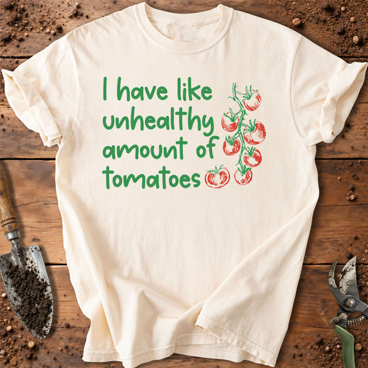 An off-white short-sleeve t-shirt with a green and red graphic print reading 'I have like unhealthy amount of tomatoes' next to an illustration of tomatoes on the vine. The shirt is laid flat on a rustic wooden background with gardening tools like a trowel and pruning shears.