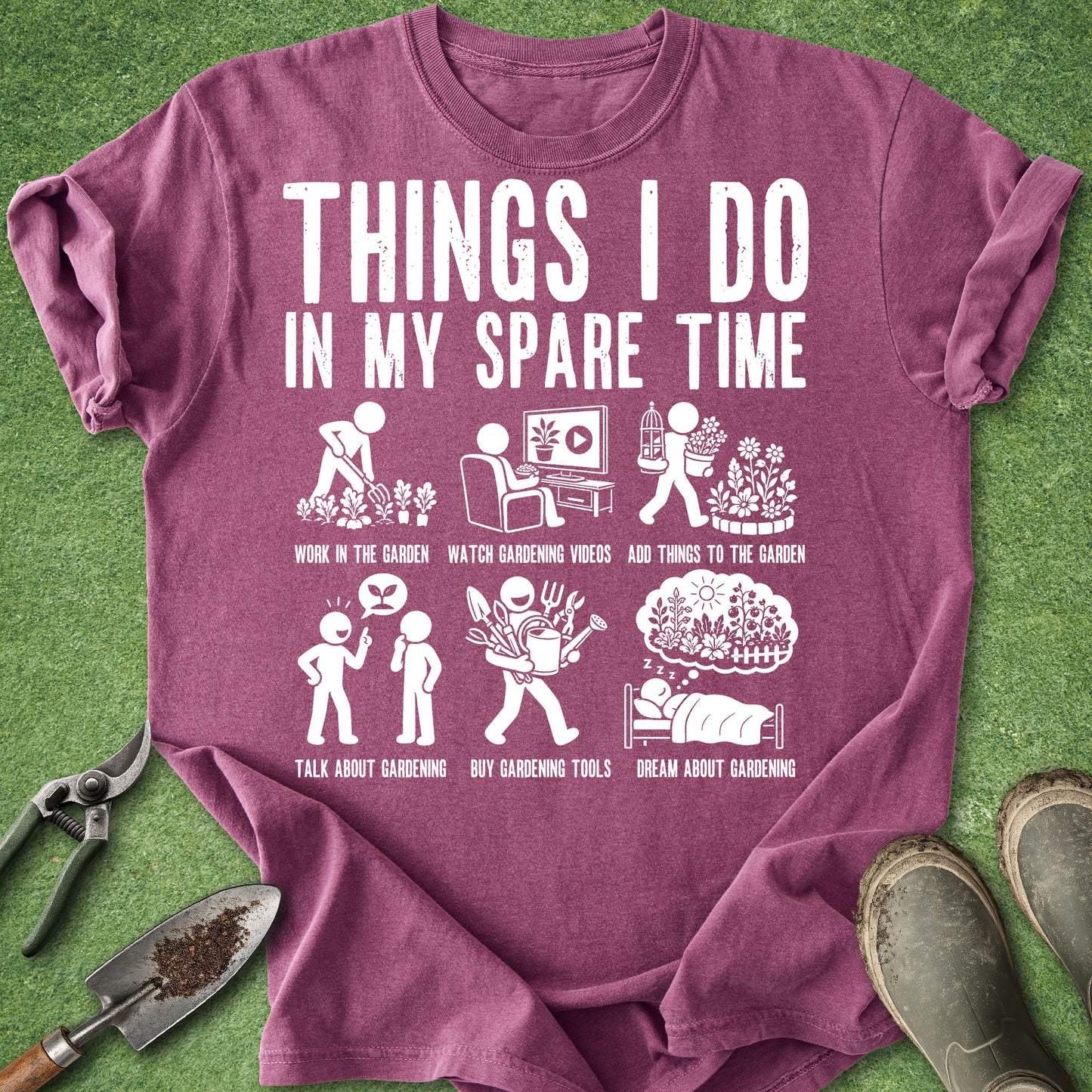 A heathered purple short-sleeve t-shirt with a white graphic print listing various gardening activities, laid flat on a green grass background with gardening props like a trowel, pruning shears, and boots.
