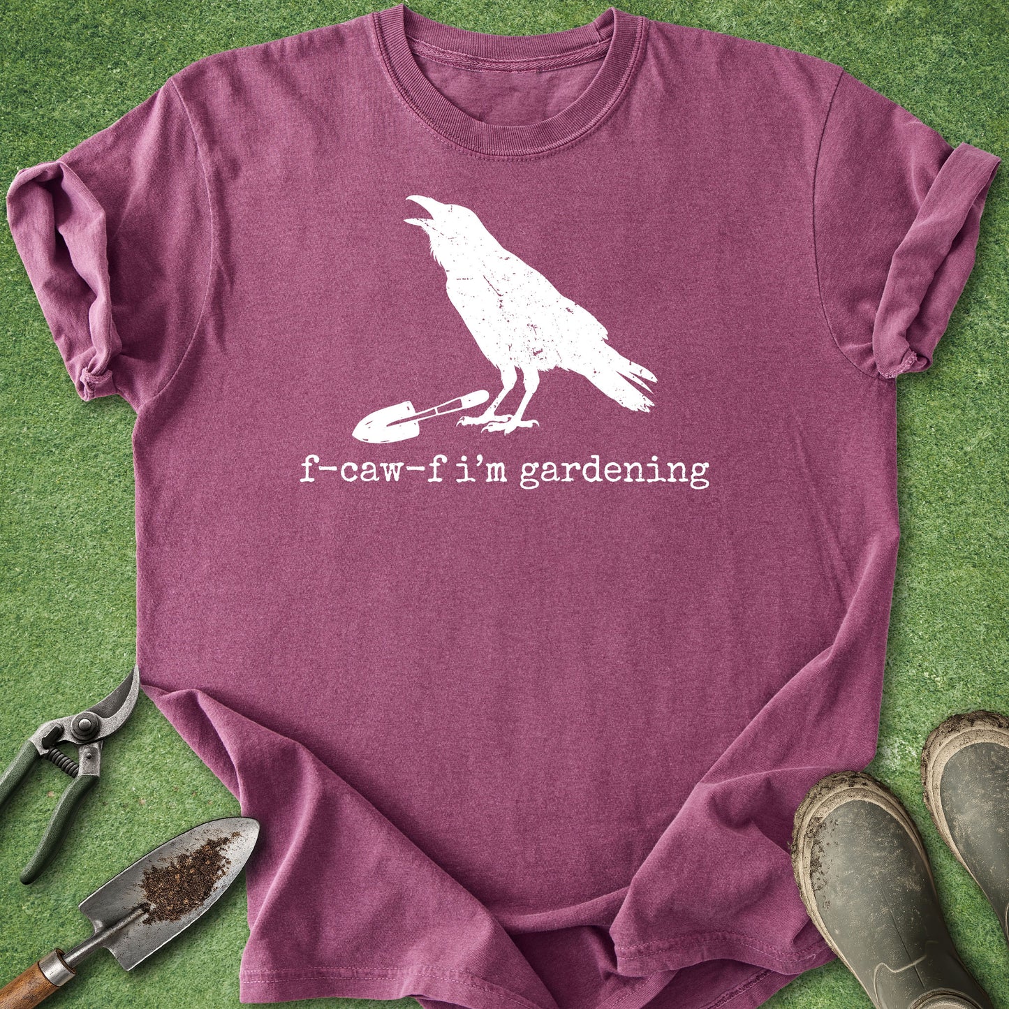 A mauve-purple short-sleeve t-shirt with a white graphic of a crow holding a gardening tool and the text 'f-caw-f i'm gardening', laid flat on green grass with gardening props including pruning shears, a trowel, and boots.