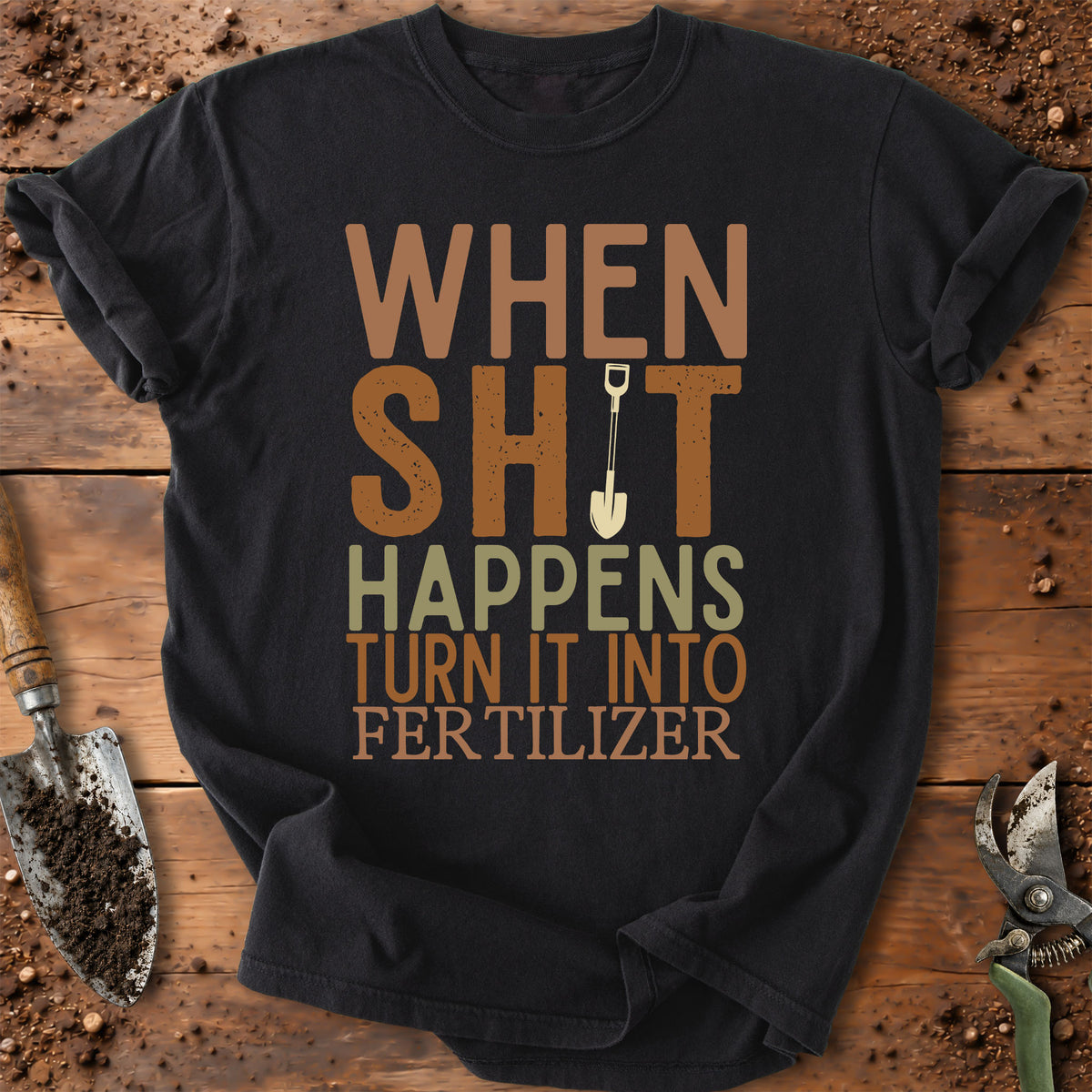A black graphic t-shirt laid flat on a rustic wooden surface with gardening props like a trowel and pruning shears. The shirt features a humorous graphic that reads 'WHEN SHIT HAPPENS TURN IT INTO FERTILIZER' in brown, green, and beige lettering, with a small shovel icon replacing the 'I' in 'SHIT'.