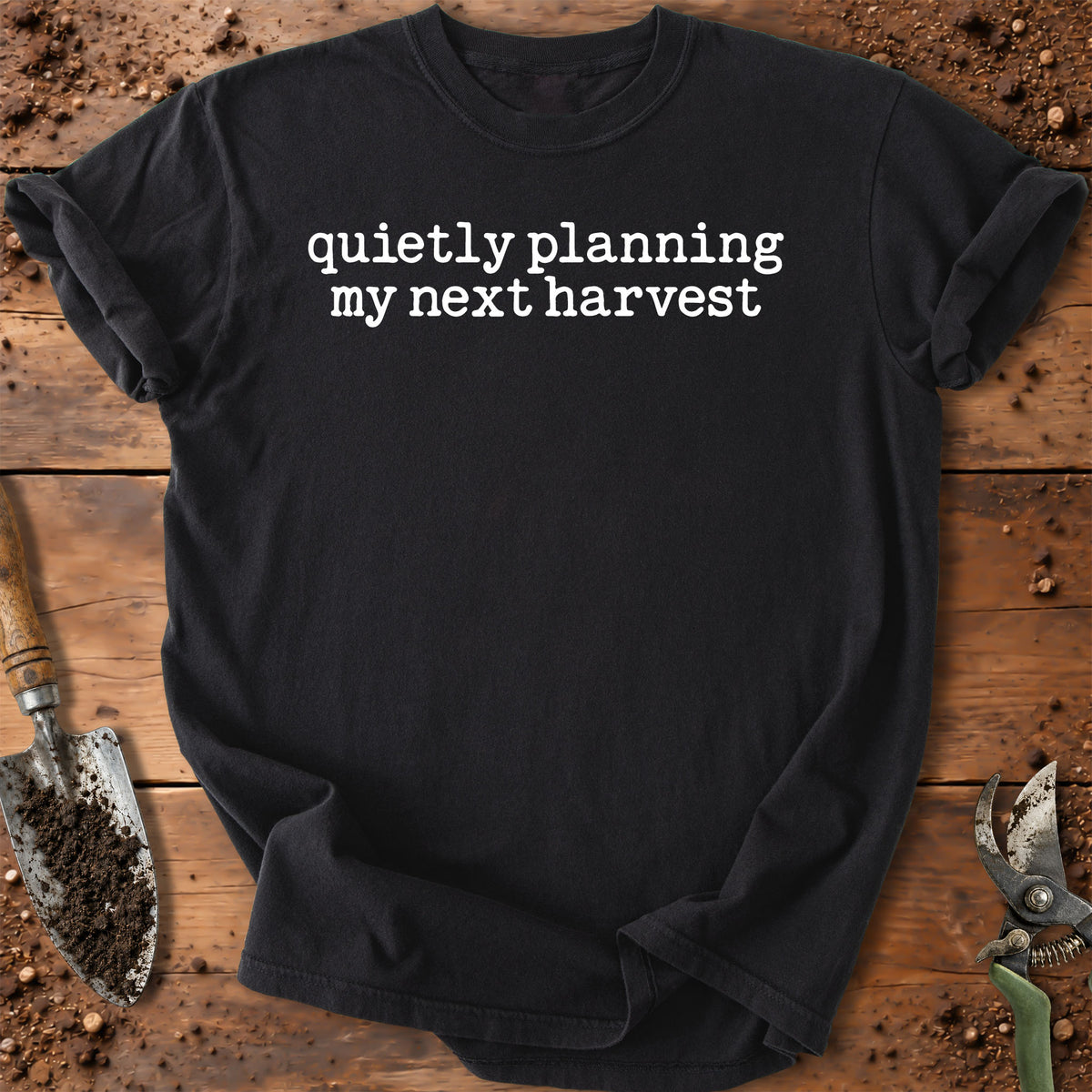 A black short-sleeve crewneck t-shirt with the white text 'quietly planning my next harvest' printed across the chest. The shirt is laid flat on a rustic wooden surface surrounded by gardening props, including a trowel and pruning shears.