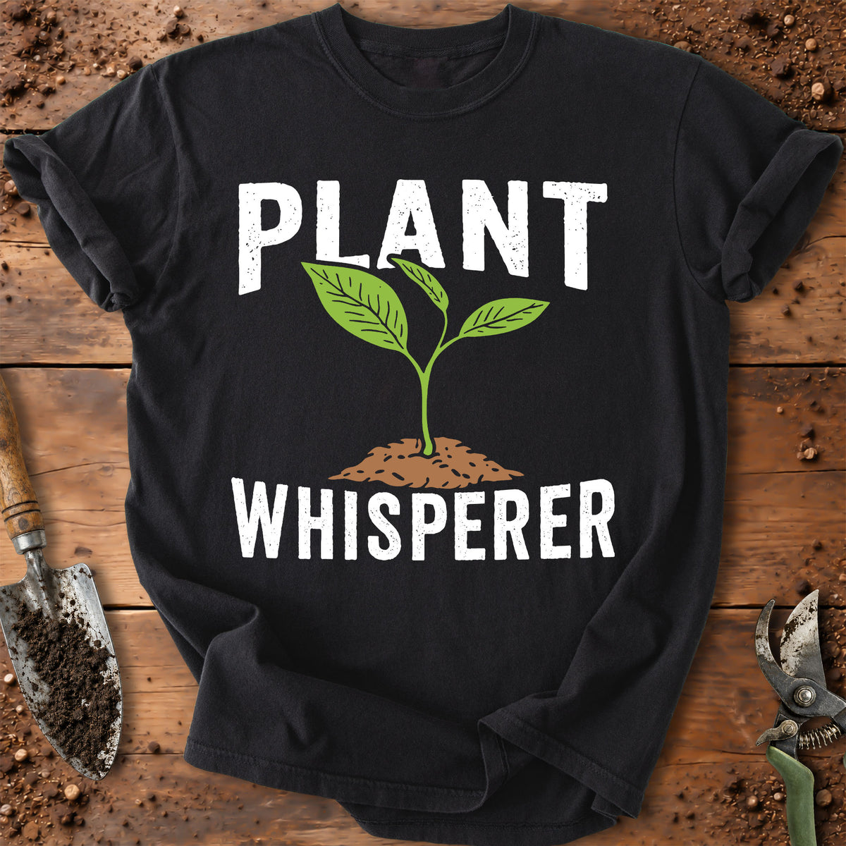 A black short-sleeve graphic T-shirt laid flat on a rustic wooden surface with soil and gardening tools around it. The shirt has large white text reading 'PLANT WHISPERER' with a green sprout and brown soil mound graphic in the center.