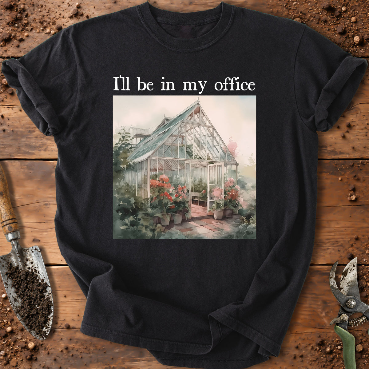 A black short-sleeve graphic T-shirt laid flat on a rustic wooden surface with gardening props. The shirt has white text reading 'I'll be in my office' above a rectangular printed illustration of a greenhouse surrounded by flowers.