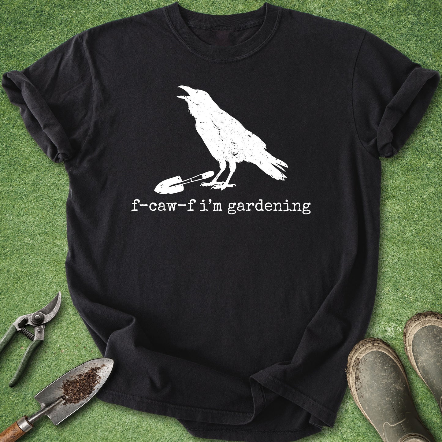 A black short-sleeve graphic T-shirt laid flat on green artificial grass, with a white raven standing on a small shovel and the text 'f-caw-f i'm gardening' printed below. Gardening props including pruning shears, a trowel with soil, and muddy boots are placed around the shirt.
