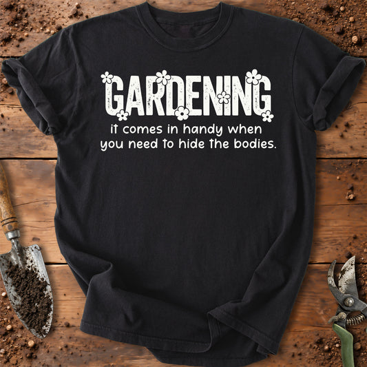 A black short-sleeve t-shirt with a white graphic text that reads 'GARDENING it comes in handy when you need to hide the bodies.' styled flat on a rustic wooden background with gardening tools like a trowel and pruning shears.