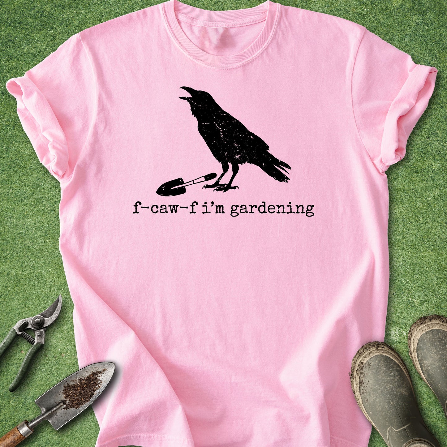 A pink short-sleeve t-shirt laid flat on green grass, featuring a black graphic of a crow holding a small shovel and the text 'f-caw-f i'm gardening'. Gardening props including a trowel, pruning shears, and boots are arranged around the shirt.