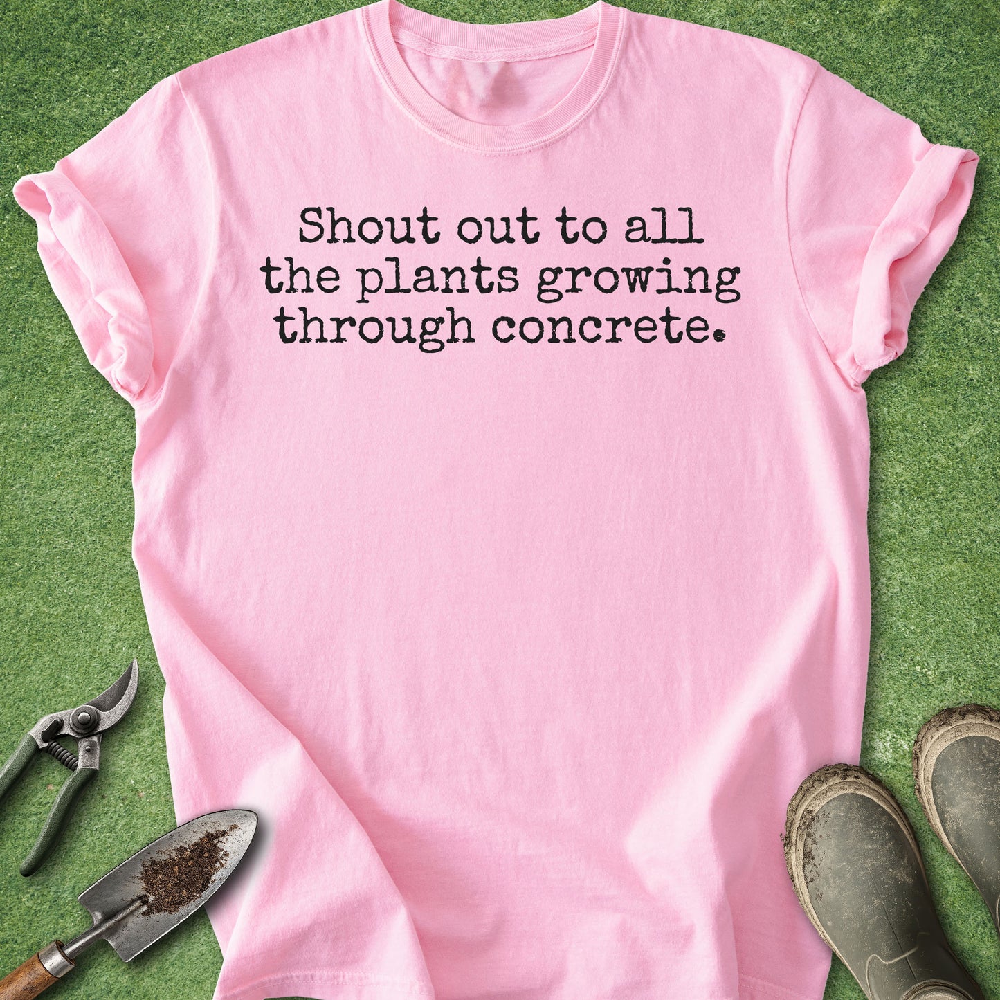 A light pink short-sleeve t-shirt with black text reading 'Shout out to all the plants growing through concrete.' laid flat on green grass with gardening tools and boots around it.