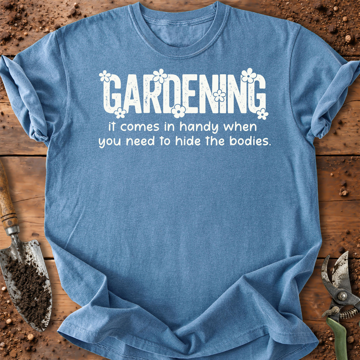 A light blue short-sleeve t-shirt with a white graphic text that reads 'GARDENING it comes in handy when you need to hide the bodies.' styled flat on a rustic wooden surface with gardening props like a trowel and pruning shears.