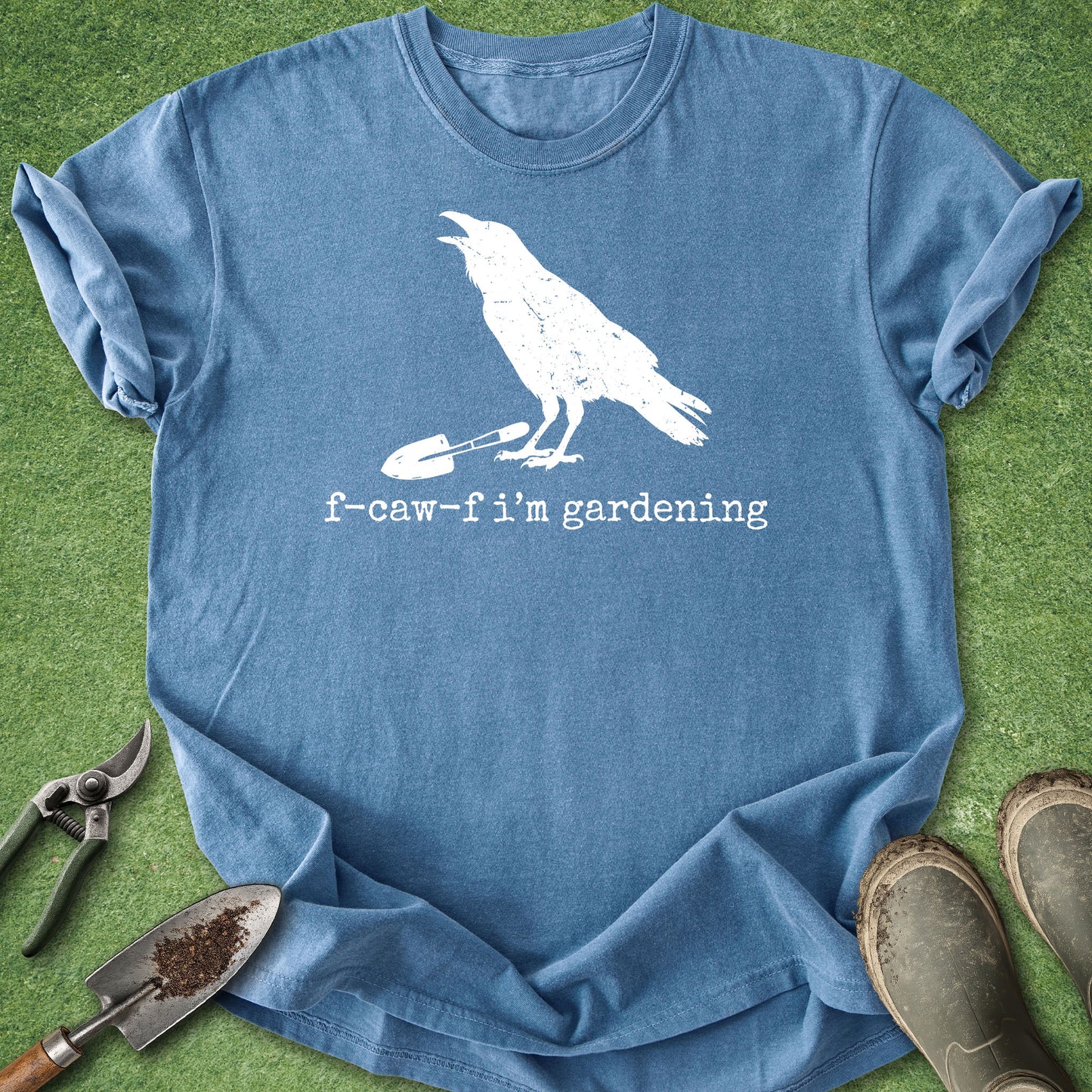 A blue short-sleeve t-shirt laid flat on green grass, featuring a white graphic of a bird holding a small shovel with the text 'f-caw-f i'm gardening'. Gardening props including a trowel, pruning shears, and boots are arranged around the shirt.