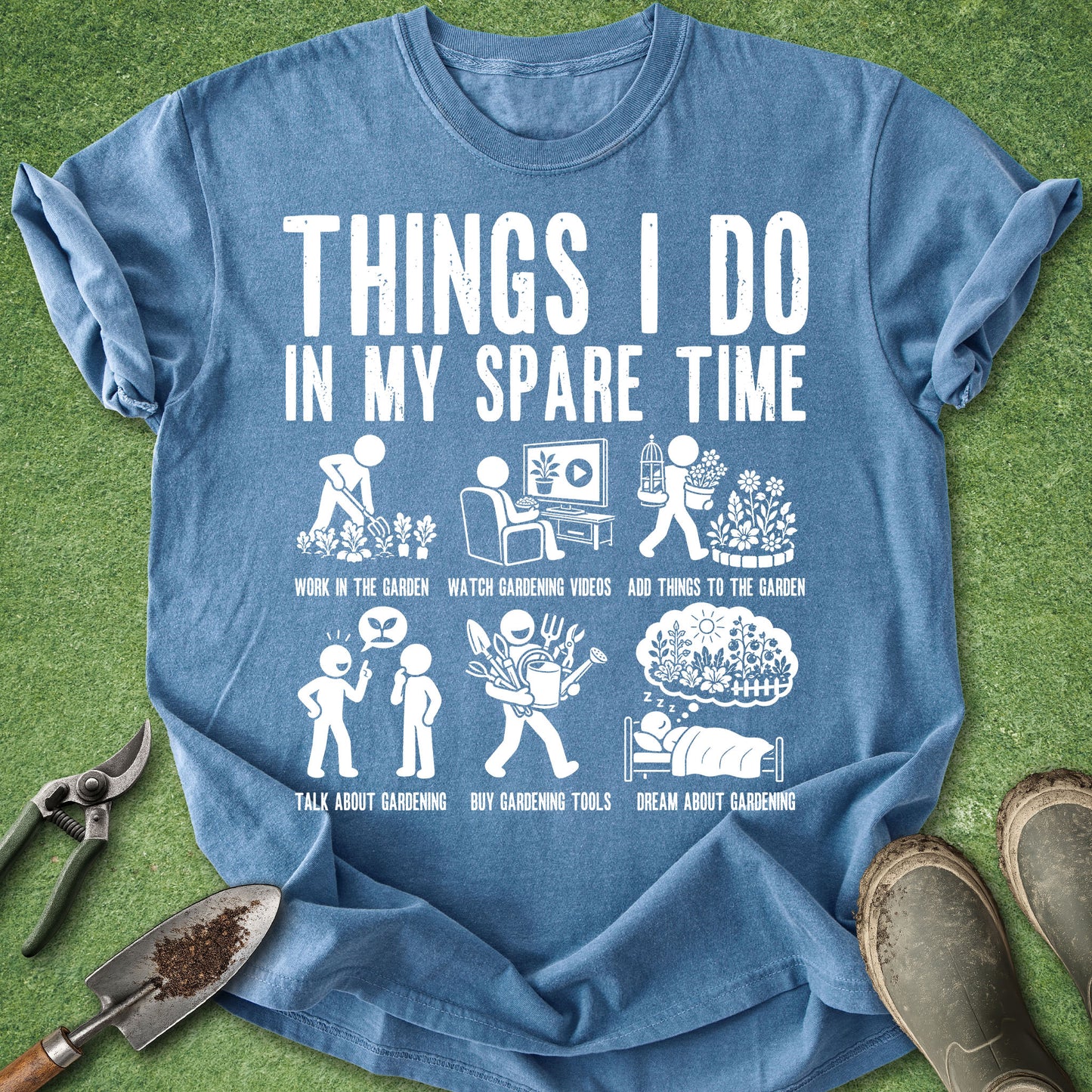 A blue graphic t-shirt laid flat on green grass with gardening props like pruning shears, a trowel, and boots around it. The shirt features a white printed graphic with the text 'THINGS I DO IN MY SPARE TIME' and six small gardening-themed illustrations and captions.
