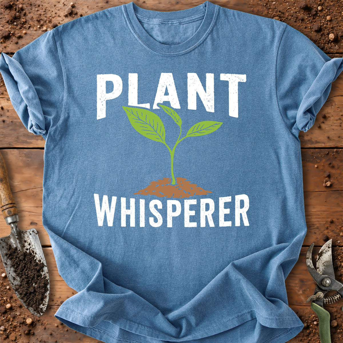 A blue short-sleeve t-shirt laid flat on a rustic wooden surface with gardening props. The shirt features a graphic print of a green sprout growing from brown soil, with the words 'PLANT WHISPERER' in large white distressed lettering.