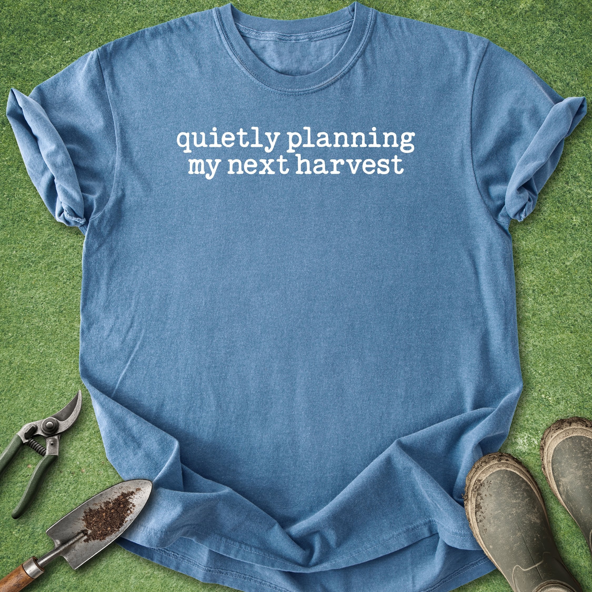 Blue t-shirt with 'quietly planning my next harvest' text on a grass background with gardening tools.