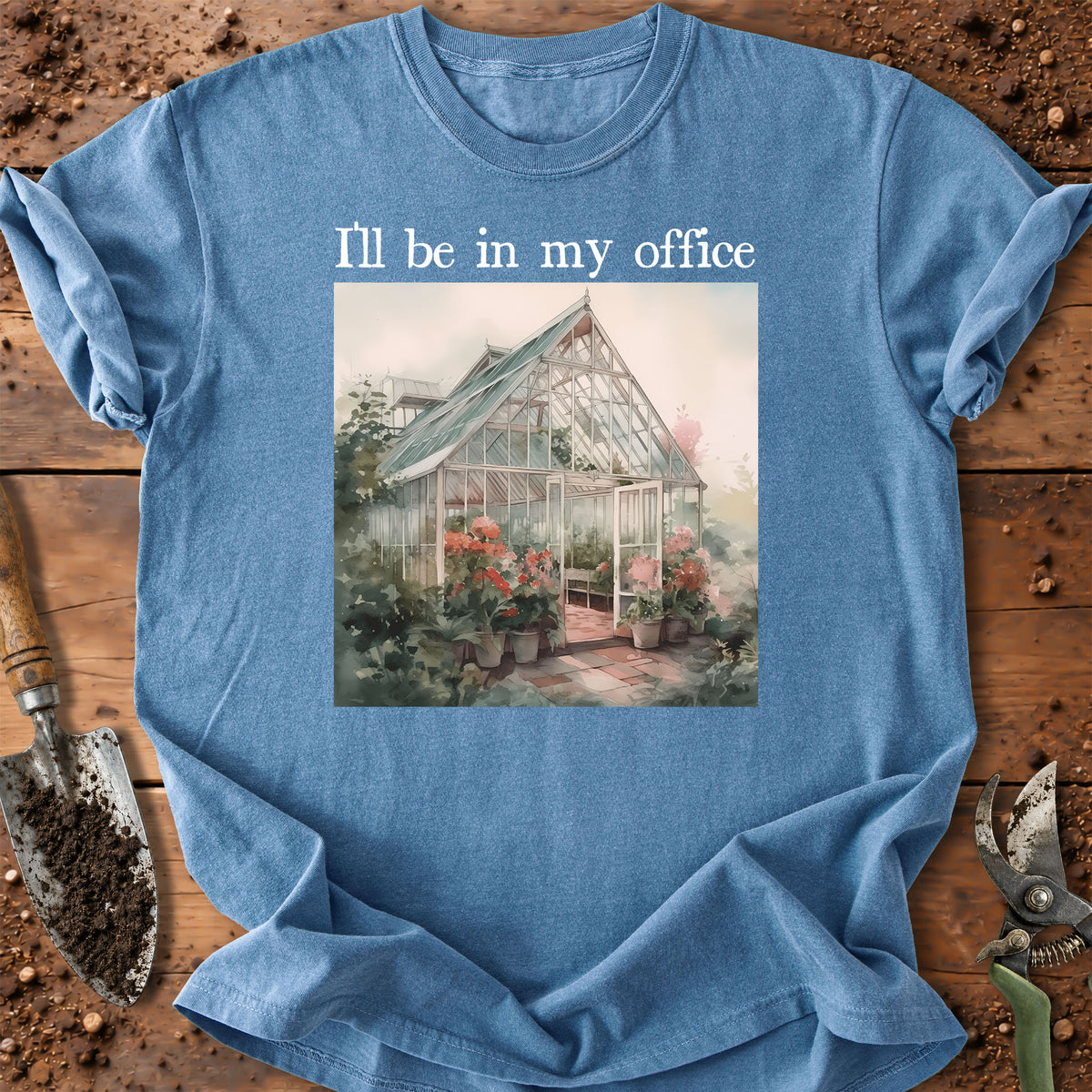A light blue graphic t-shirt laid flat on a rustic wooden surface with gardening tools. The shirt features the text 'I'll be in my office' above a vintage-style illustration of a greenhouse surrounded by flowers.
