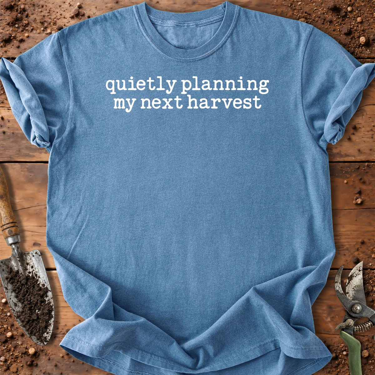 A light blue crewneck t-shirt with the phrase 'quietly planning my next harvest' printed in white lowercase serif text across the chest. The shirt is laid flat on a rustic wooden surface with gardening tools like a trowel and pruning shears visible around the edges.