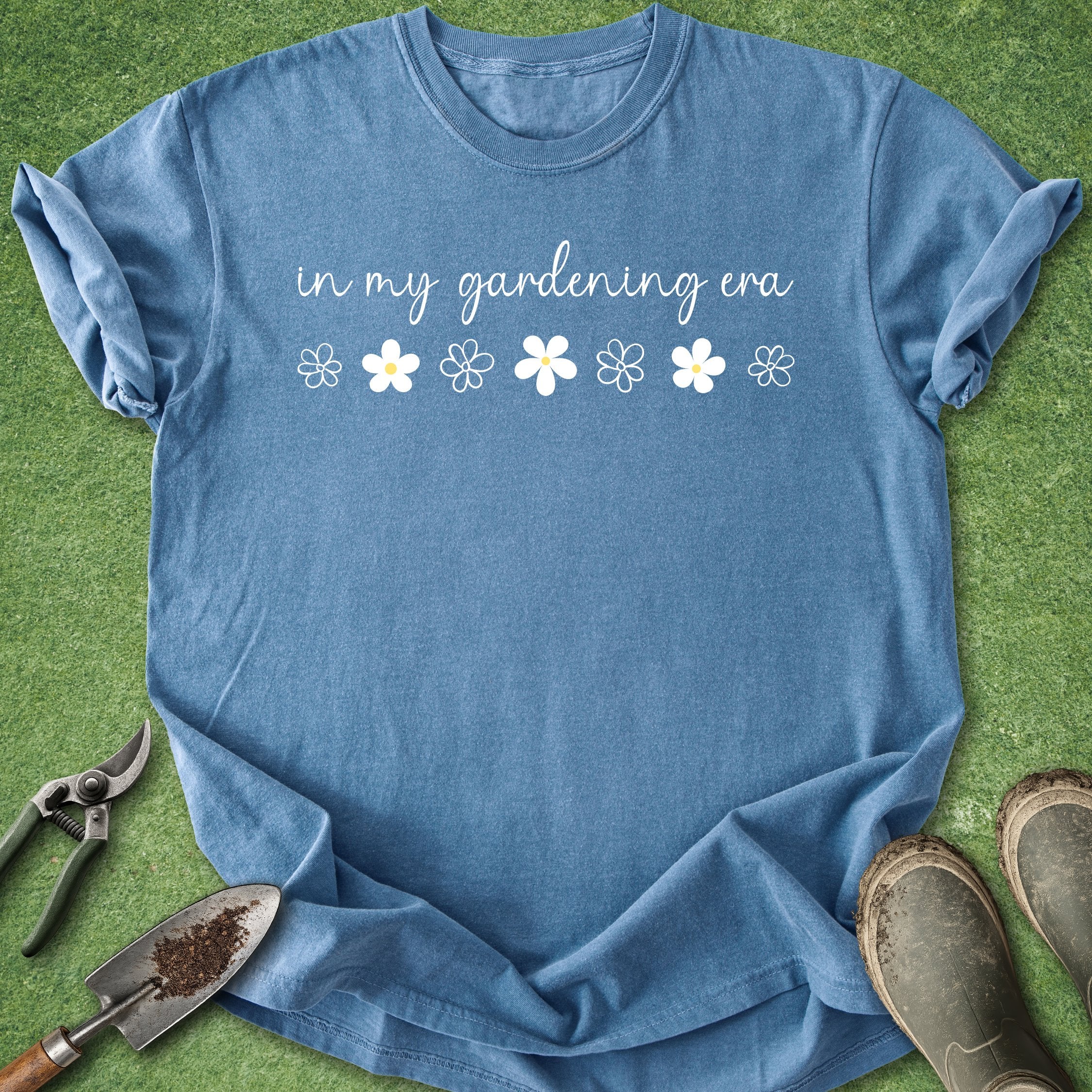 Blue t-shirt with 'in my gardening era' text and flower graphics on a grass background
