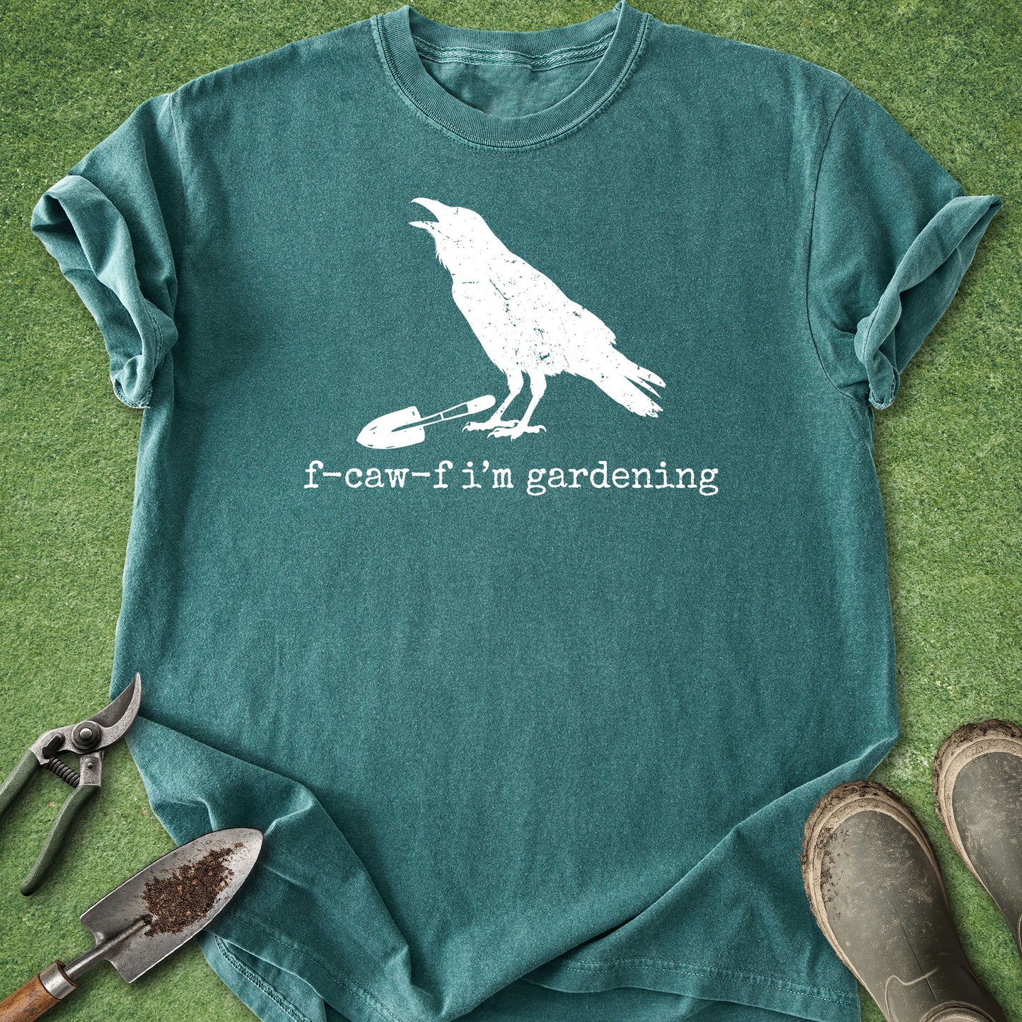 A teal t-shirt with a white graphic of a crow holding a garden fork and the text 'f-caw-f i'm gardening', laid flat on green grass with gardening props including shears, a trowel, and boots.