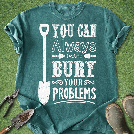 A teal graphic t-shirt laid flat on green grass with gardening props, featuring the white distressed text 'YOU CAN Always BURY YOUR PROBLEMS' alongside a shovel illustration.