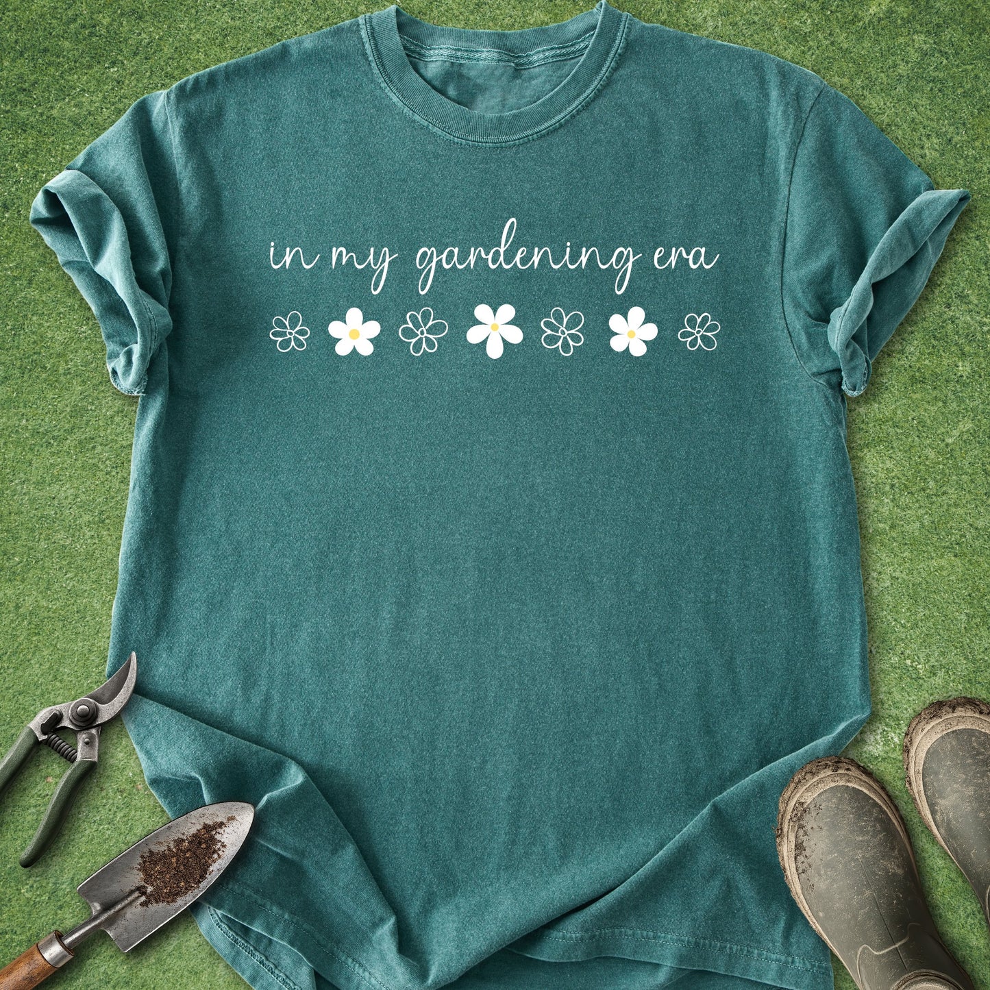 Teal t-shirt with 'in my gardening era' text and flower graphics on a grass background