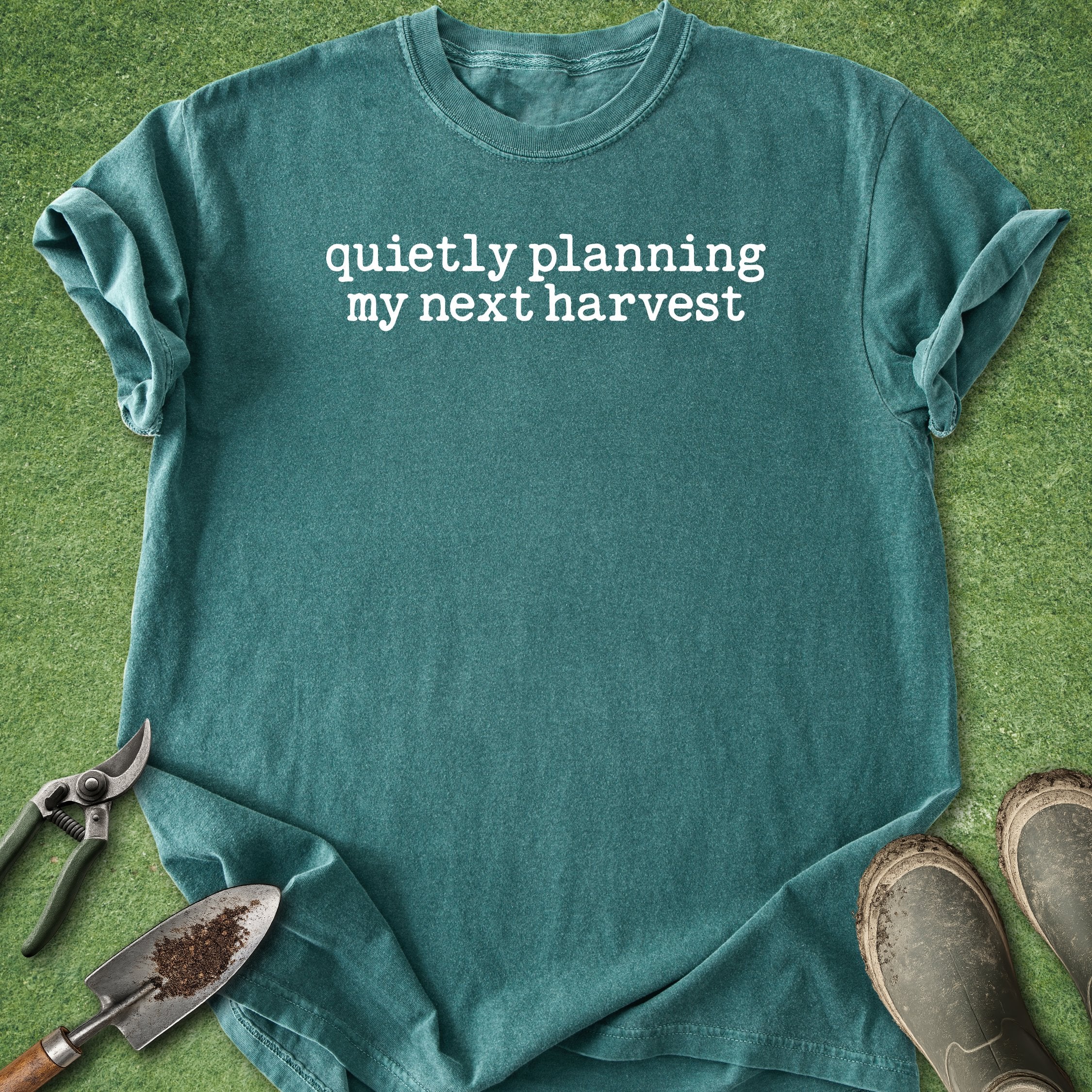 Teal t-shirt with 'quietly planning my next harvest' text on a grass background with gardening tools.