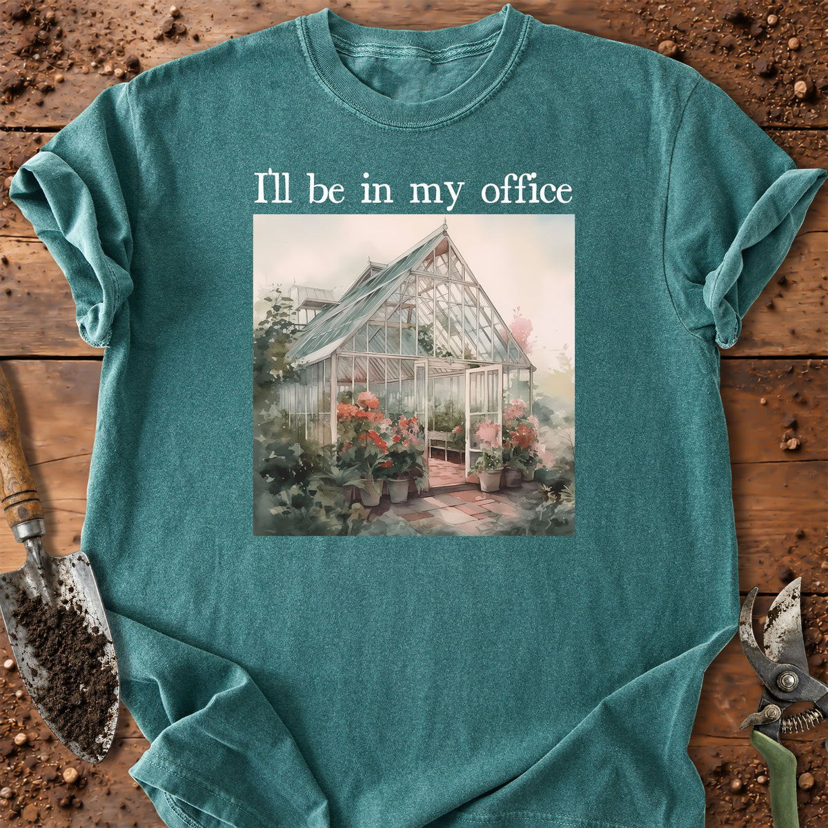A teal crewneck t-shirt laid flat on a rustic wooden surface with gardening props like a trowel and pruning shears. The shirt features a printed graphic of a greenhouse surrounded by flowers and the text 'I'll be in my office' in white.