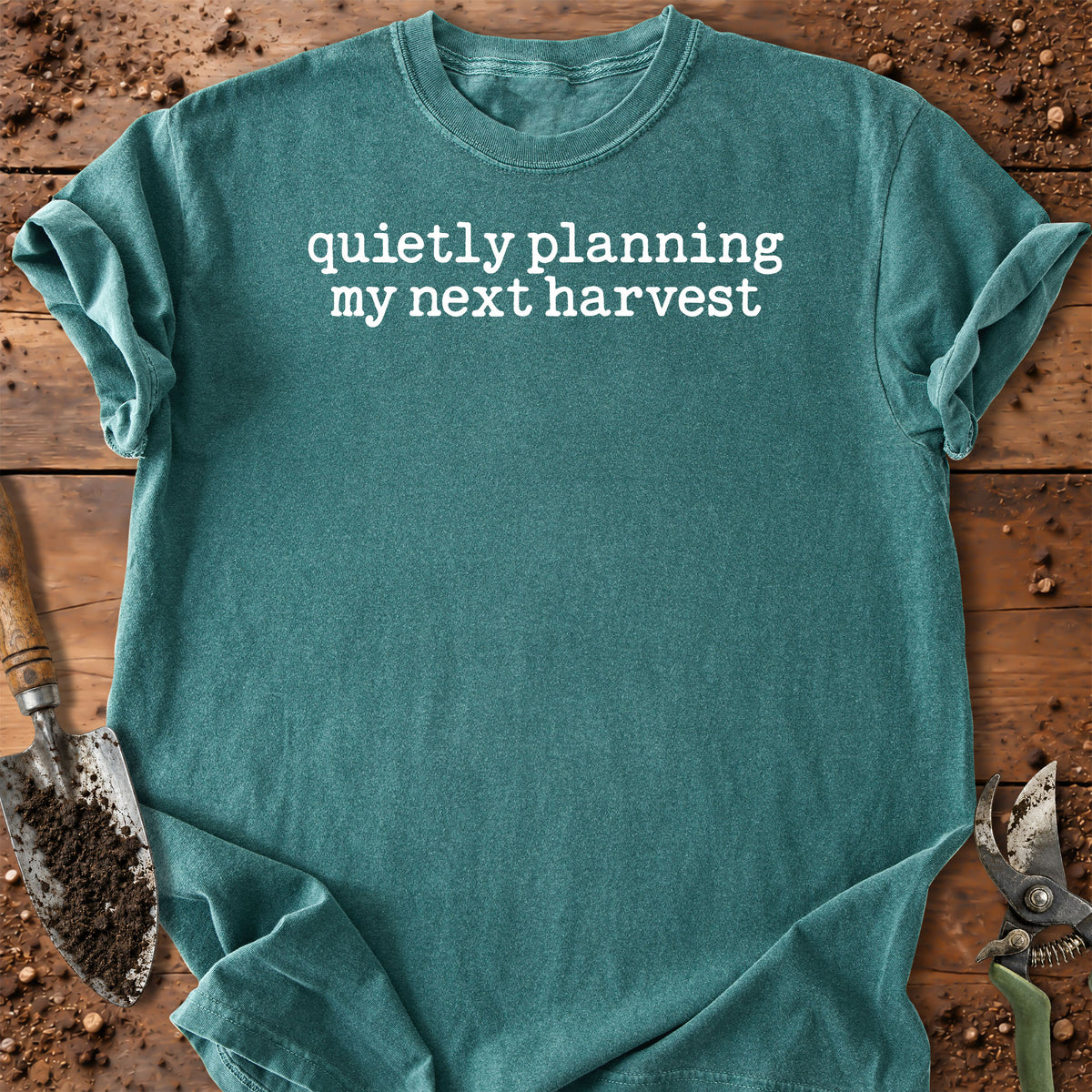A teal crewneck t-shirt with the white text 'quietly planning my next harvest' printed across the chest, laid flat on a rustic wooden surface with gardening tools like a trowel and pruning shears visible around the edges.