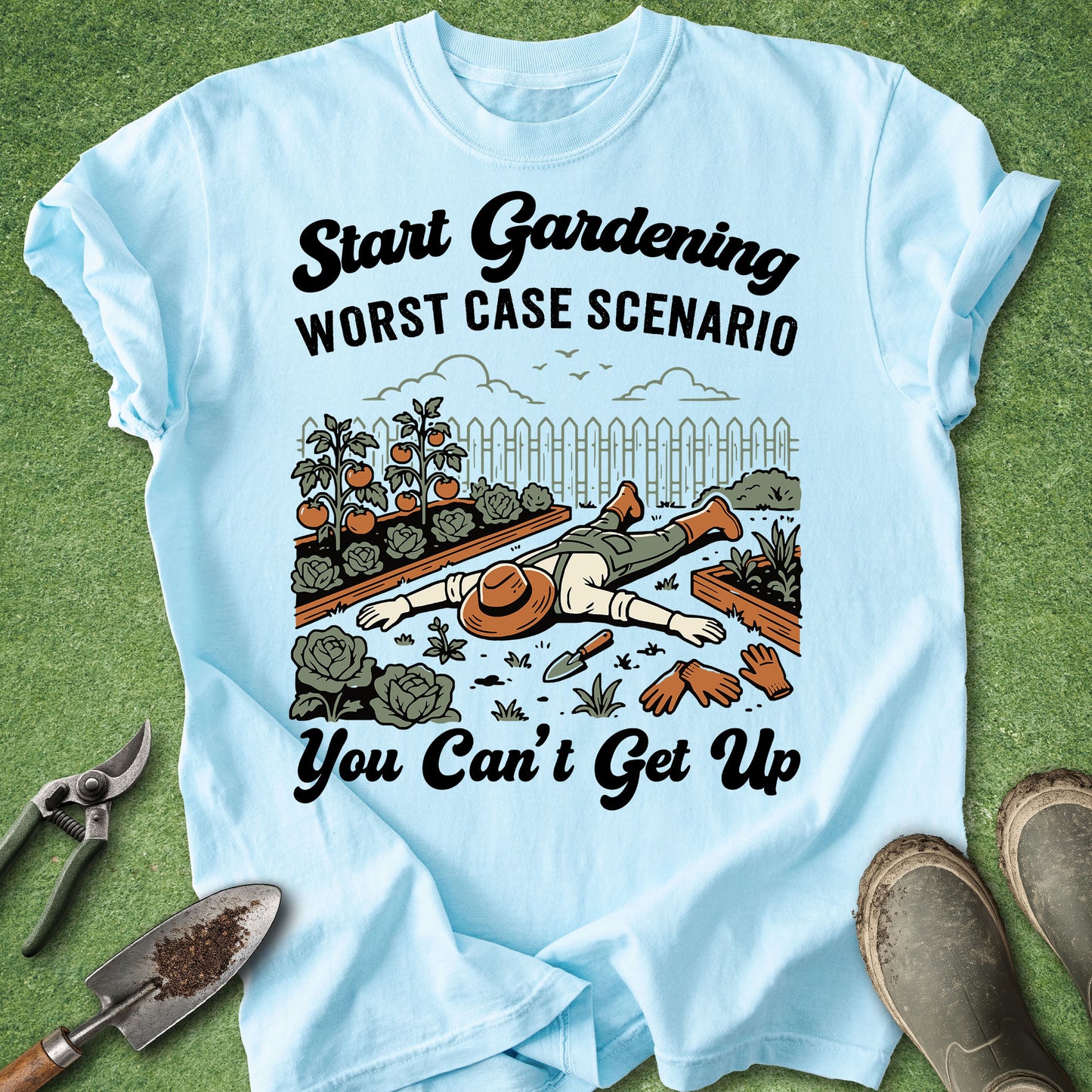 A light blue graphic t-shirt laid flat on a green grass-like surface, surrounded by gardening props including pruning shears, a trowel, and gardening boots. The shirt features a humorous illustration of a gardener lying on the ground with the text 'Start Gardening WORST CASE SCENARIO You Can't Get Up'.
