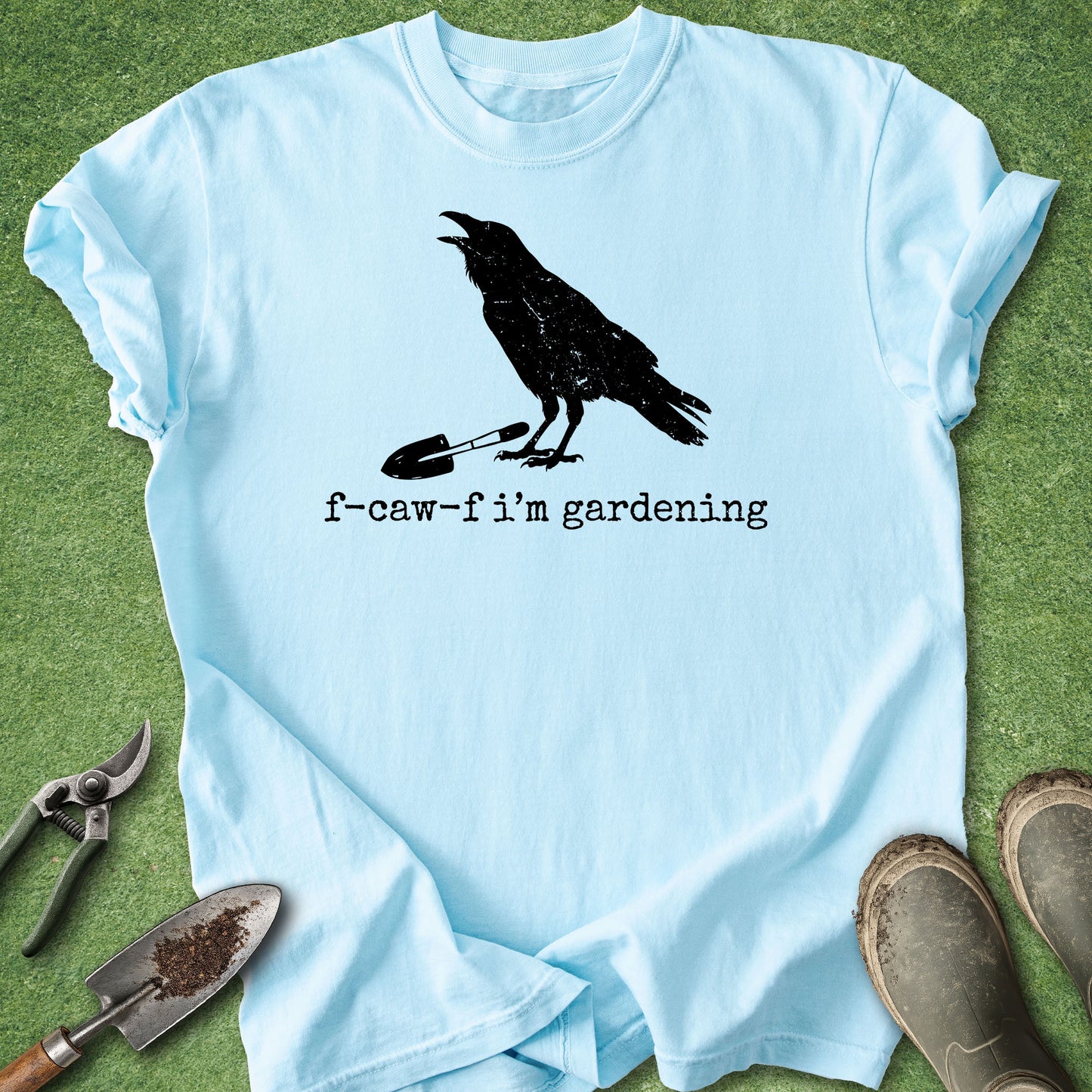 A light blue short-sleeve t-shirt with a black graphic of a crow standing on a gardening tool and the text 'f-caw-f i'm gardening', laid flat on green grass with gardening props including pruning shears, a trowel, and boots.