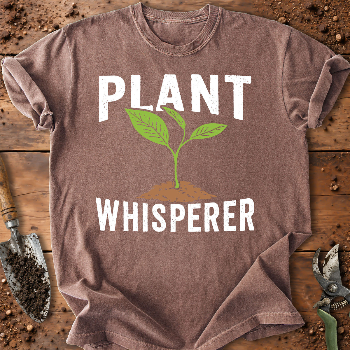 A brown short-sleeve t-shirt with a graphic that reads 'PLANT WHISPERER' in white text, featuring a green sprout growing from a brown patch of soil. The shirt is laid flat on a rustic wooden surface with gardening tools like a trowel and pruning shears visible around the edges.