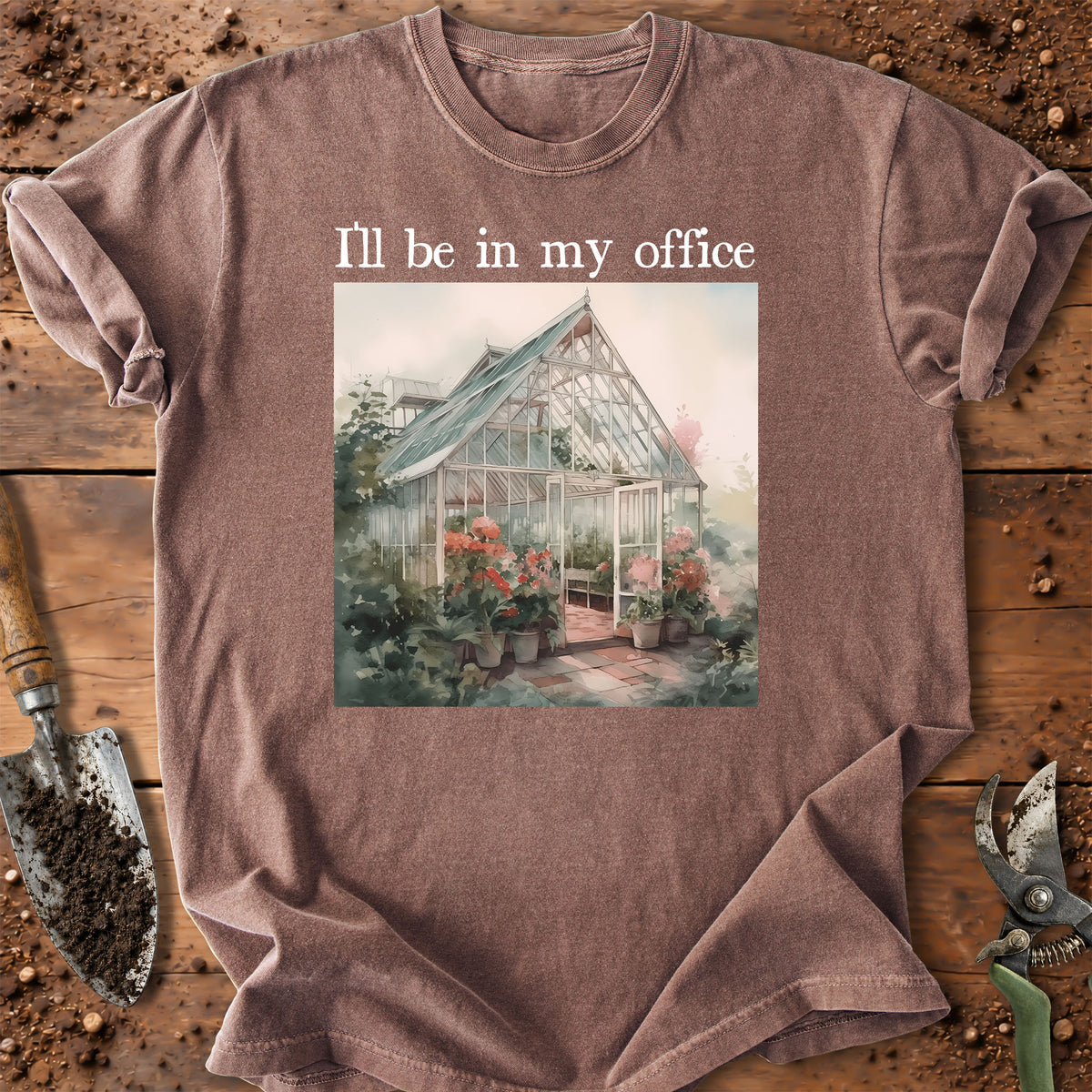 A heathered taupe crewneck t-shirt laid flat on a rustic wooden surface with gardening props. The shirt features a graphic of a greenhouse with flowers and the text 'I'll be in my office' printed above it.