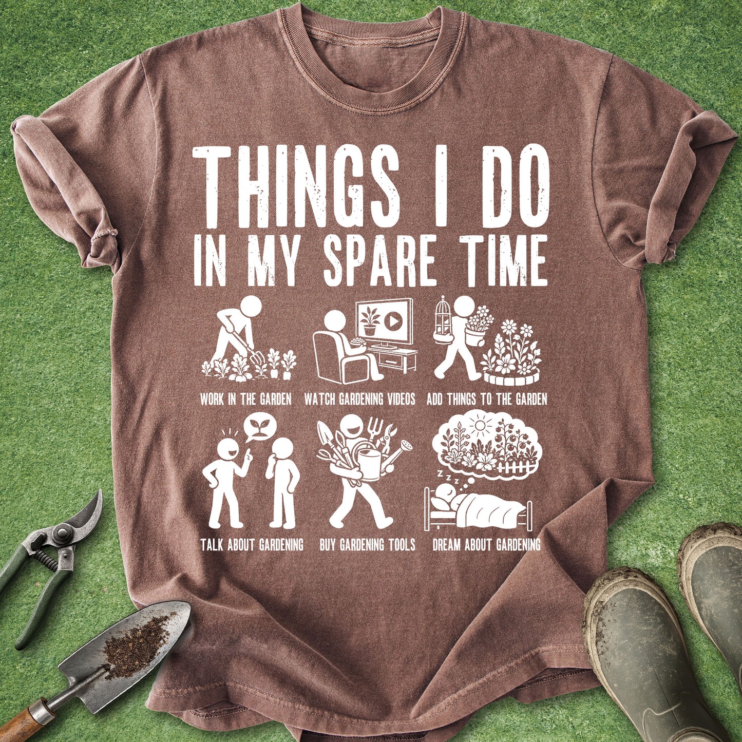 A brown short-sleeve graphic T-shirt laid flat on green grass-like background, with gardening tools and boots around it. The shirt has large white text reading 'THINGS I DO IN MY SPARE TIME' and a white illustrated list of activities such as work in the garden, watch gardening videos, add things to the garden, talk about gardening, buy gardening tools, and dream about gardening.